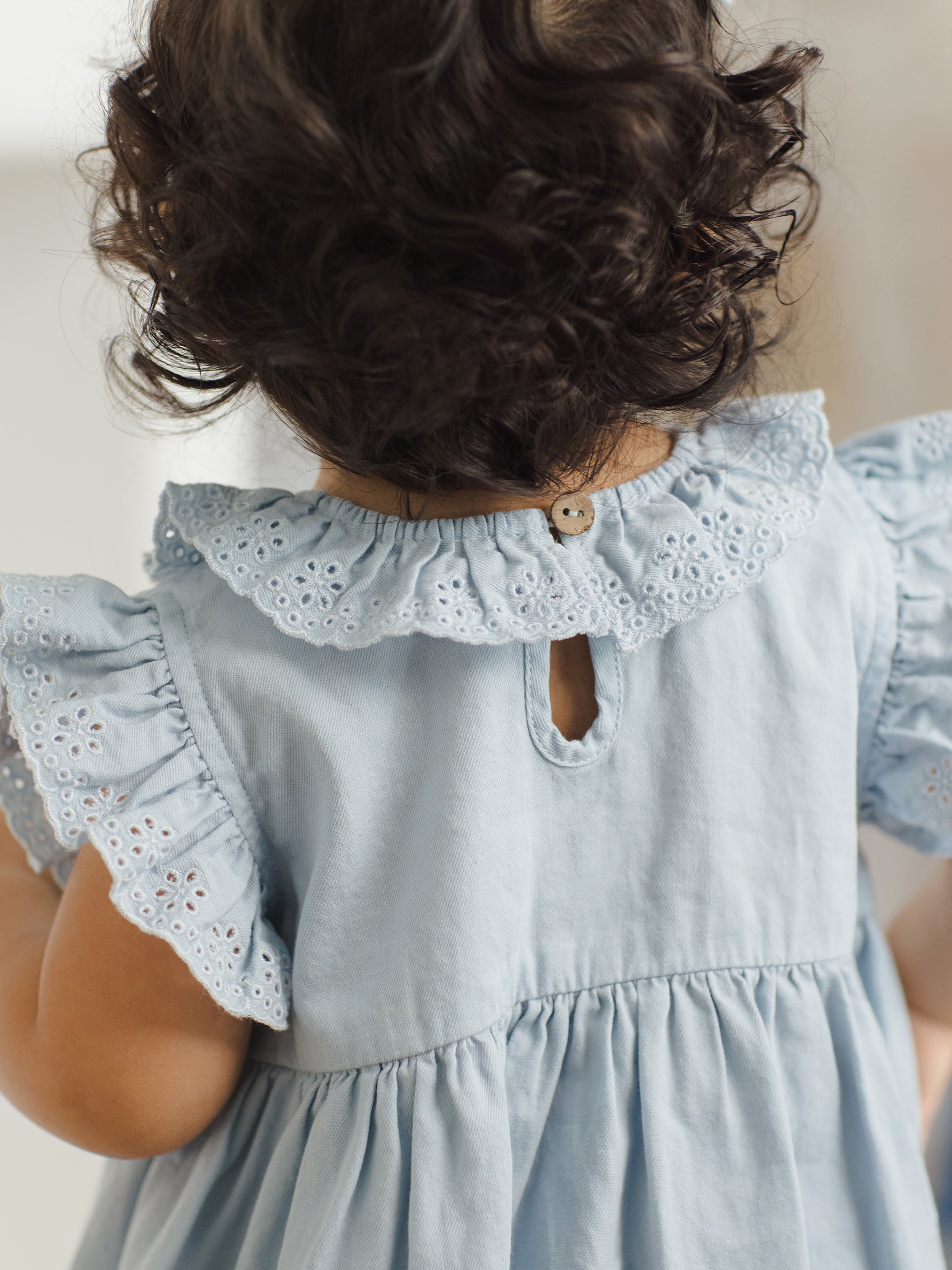 Child's back in a light blue dress with eyelet trim, showcasing curly hair and a button closure.