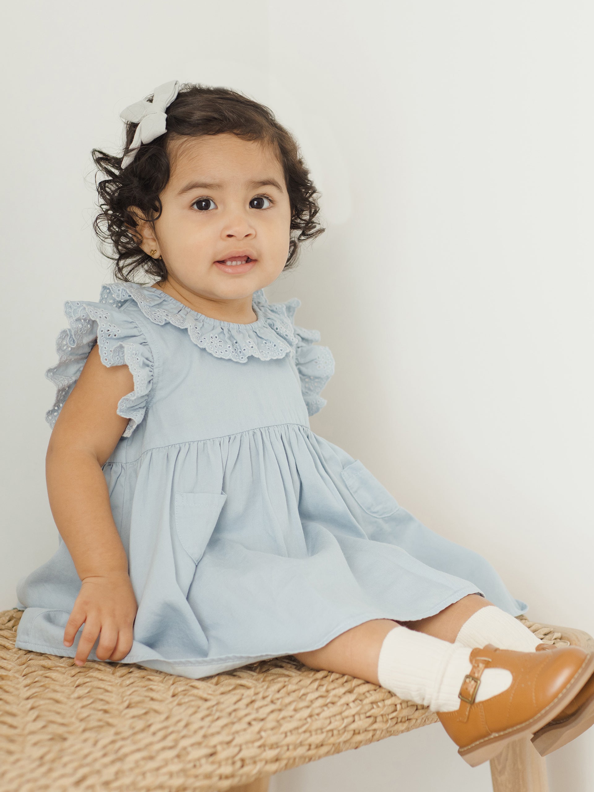 Toddler girl in a blue dress sitting on a woven stool, smiling with curly hair and a bow.