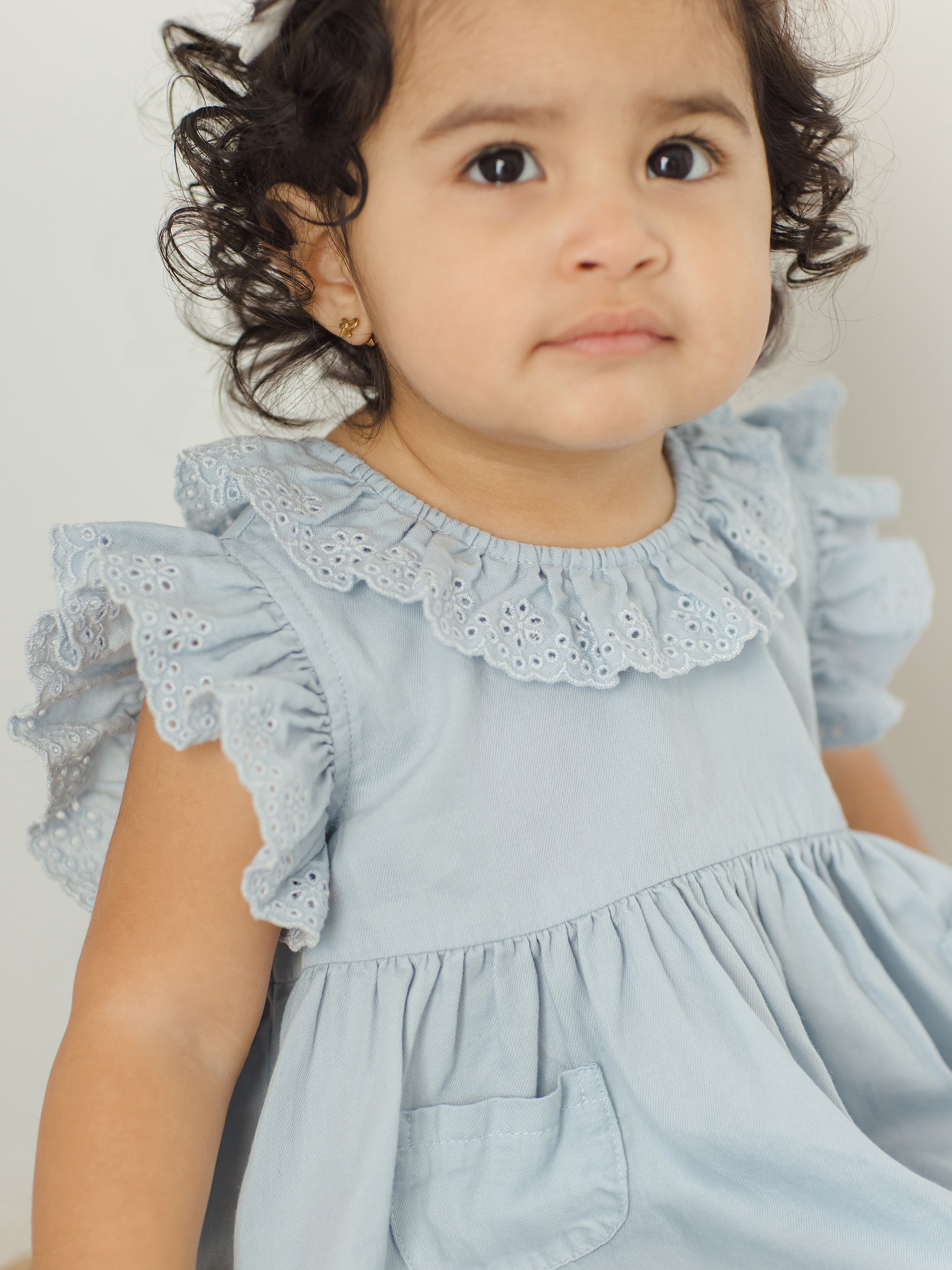 Toddler girl with curly hair wearing a light blue dress with a lace collar, looking directly at the camera.
