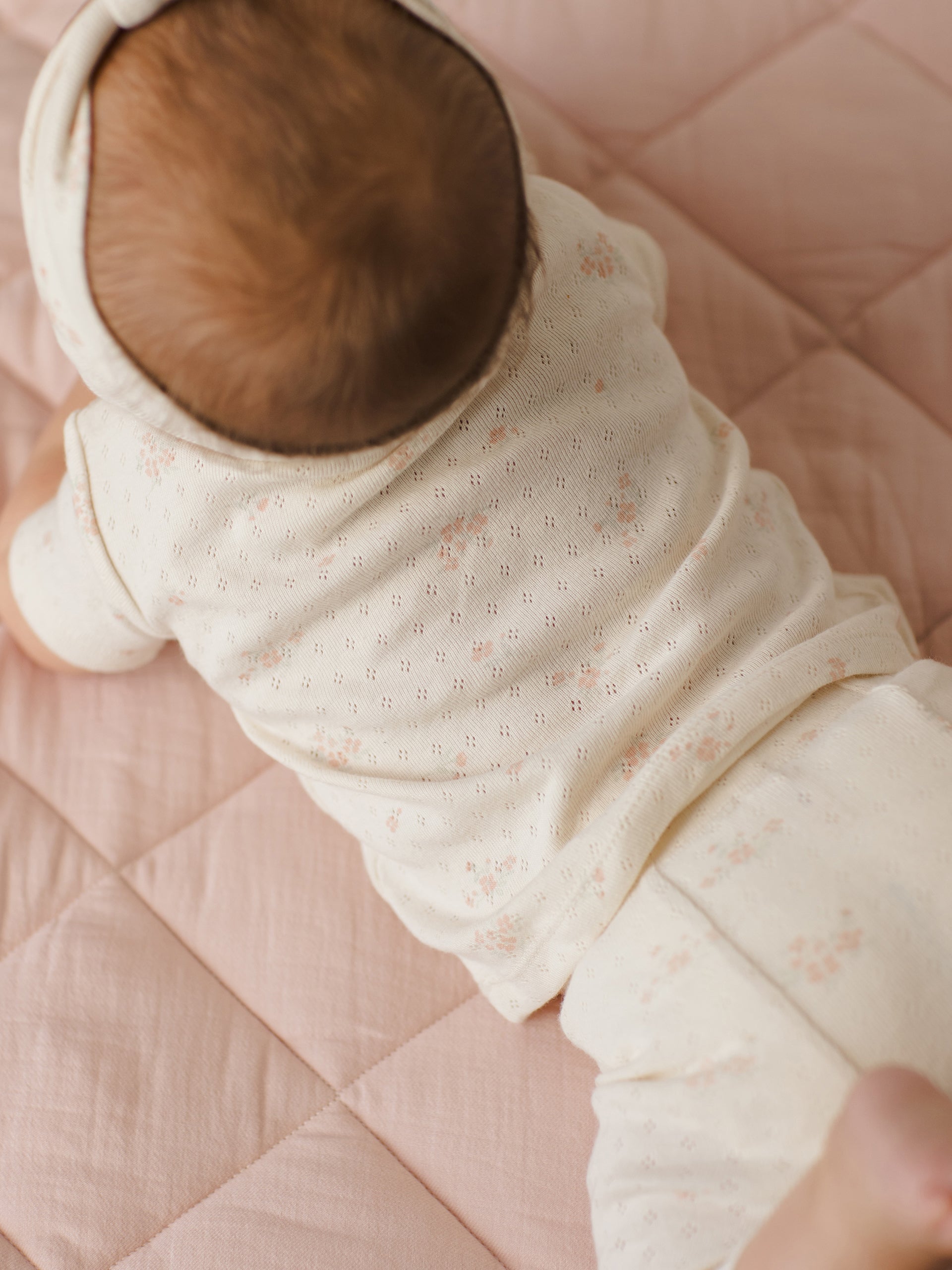 Baby crawling on a pink quilt, wearing a floral outfit and headband, showcasing soft textures and colors.
