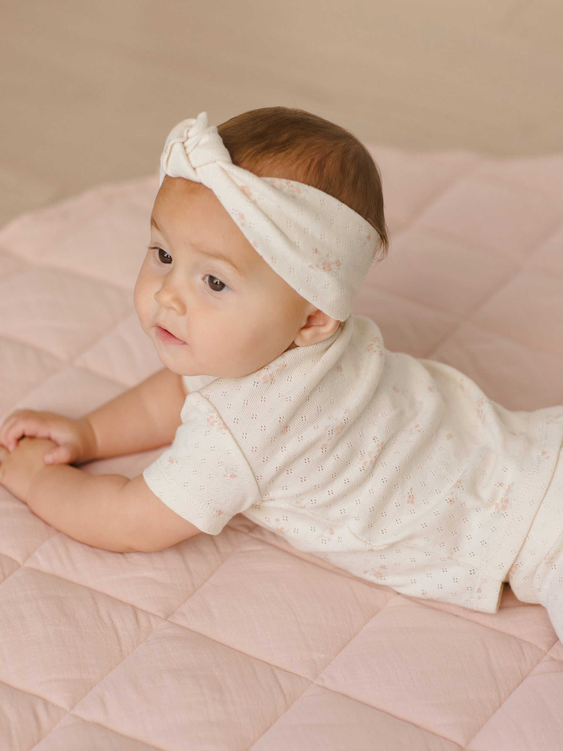 Baby with a headband lying on a pink quilted mat, looking to the side.