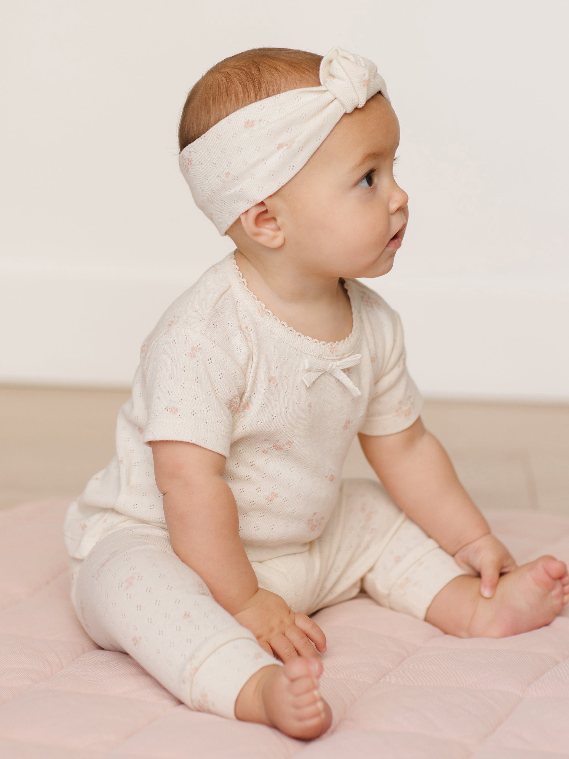 Infant girl wearing a floral outfit and headband, sitting on a pink mat, looking thoughtfully to the side.