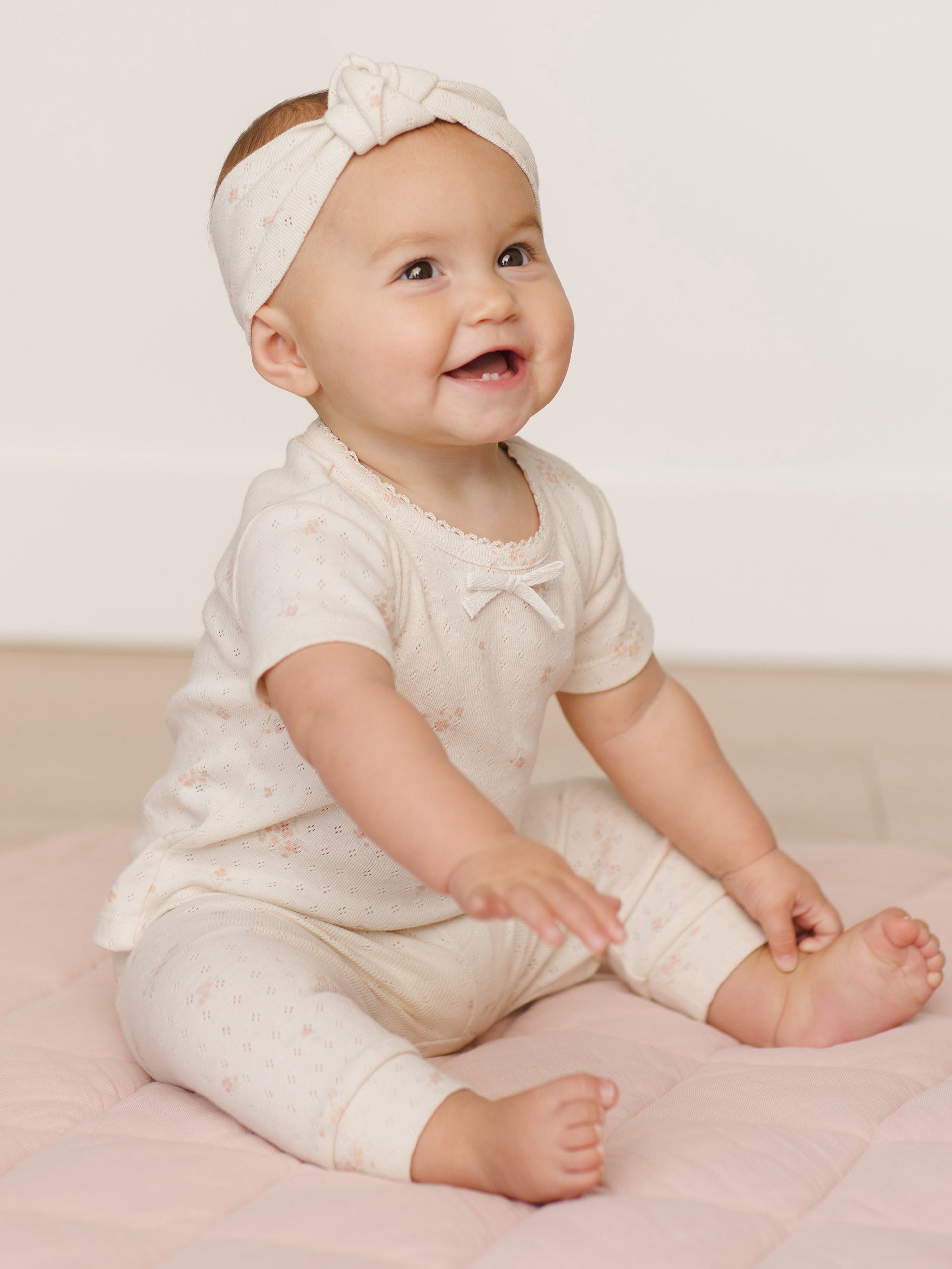Smiling baby in a cream outfit sits on a soft mat, reaching out with joy.