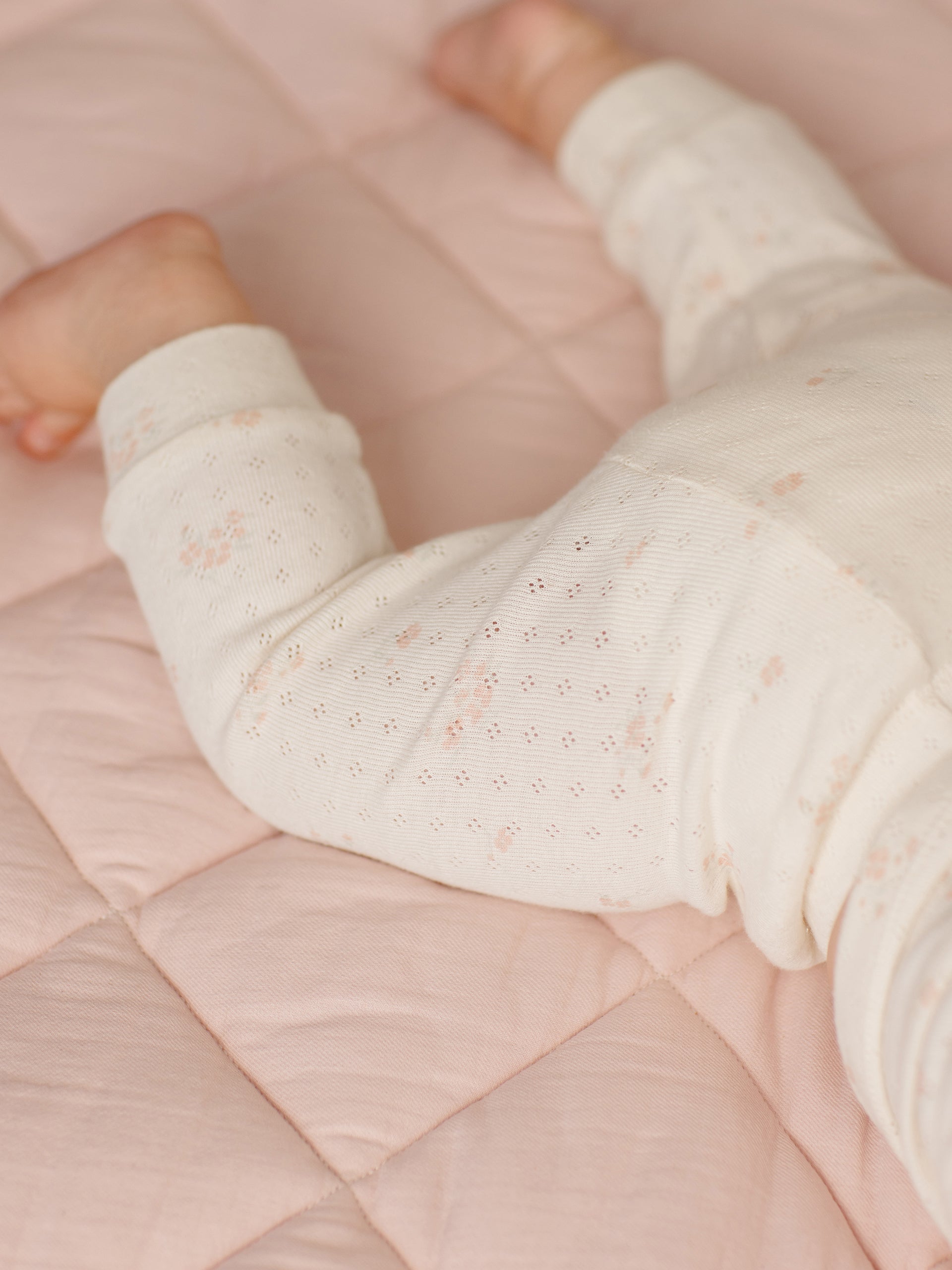 Baby's legs in cream patterned pajamas on a soft pink quilted blanket.