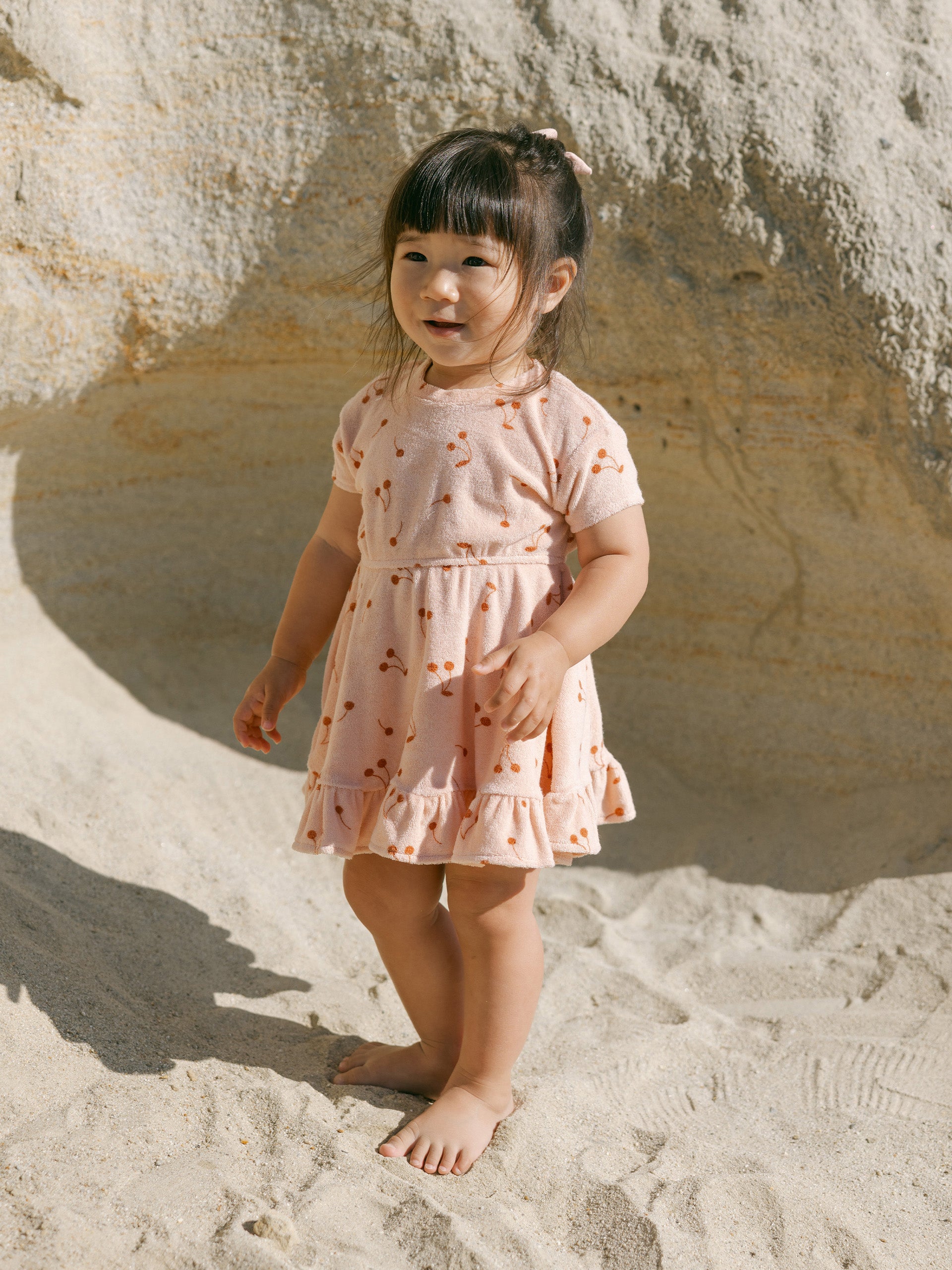 Young girl in a peach dress with cherry patterns, standing barefoot on sandy ground by a rock.