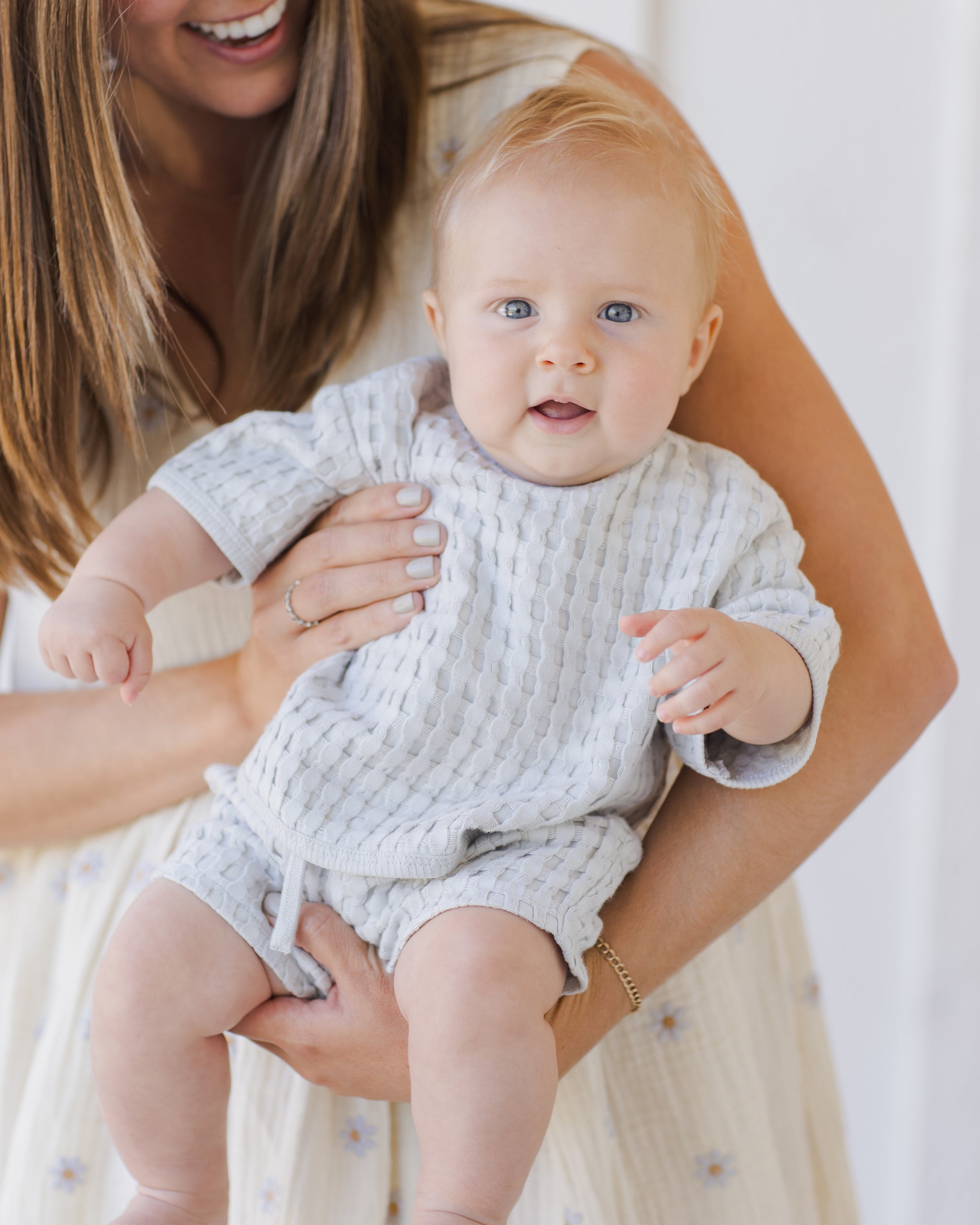 Smiling woman holds an adorable baby wearing a light gray outfit, both enjoying a warm and joyful moment together.