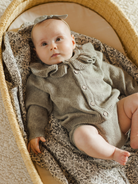 Baby in a cozy basket, wearing a knitted outfit and a bow headband, gazing upward.