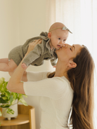 Mother kissing her baby while holding them up, surrounded by soft sunlight and green plants indoors.