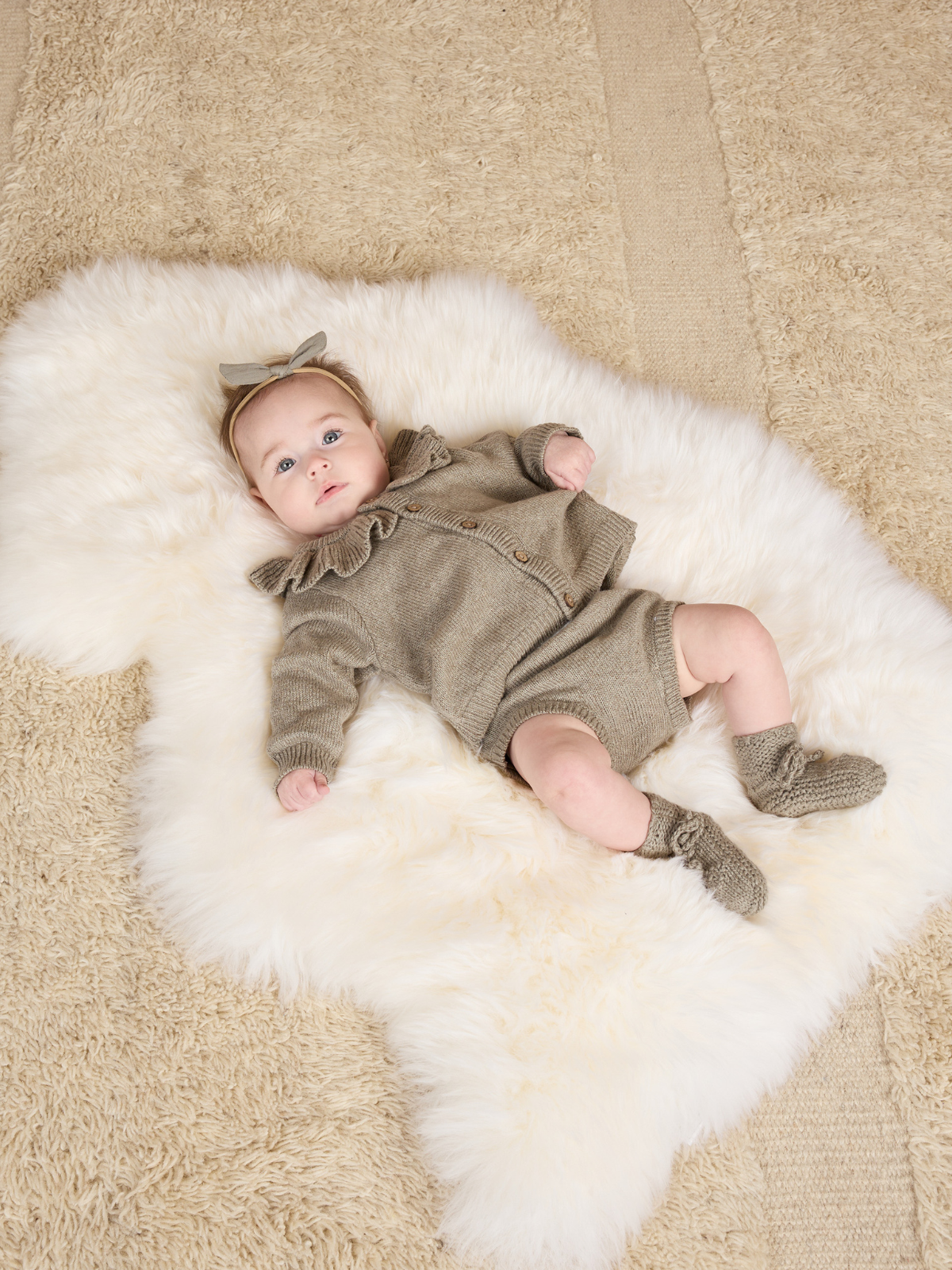 Baby girl with a bow headband, wearing a gray outfit, lying on a fluffy white rug.