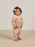 Toddler with curly hair and a bow, wearing a pink outfit, smiling in a light-filled room with wooden accents.