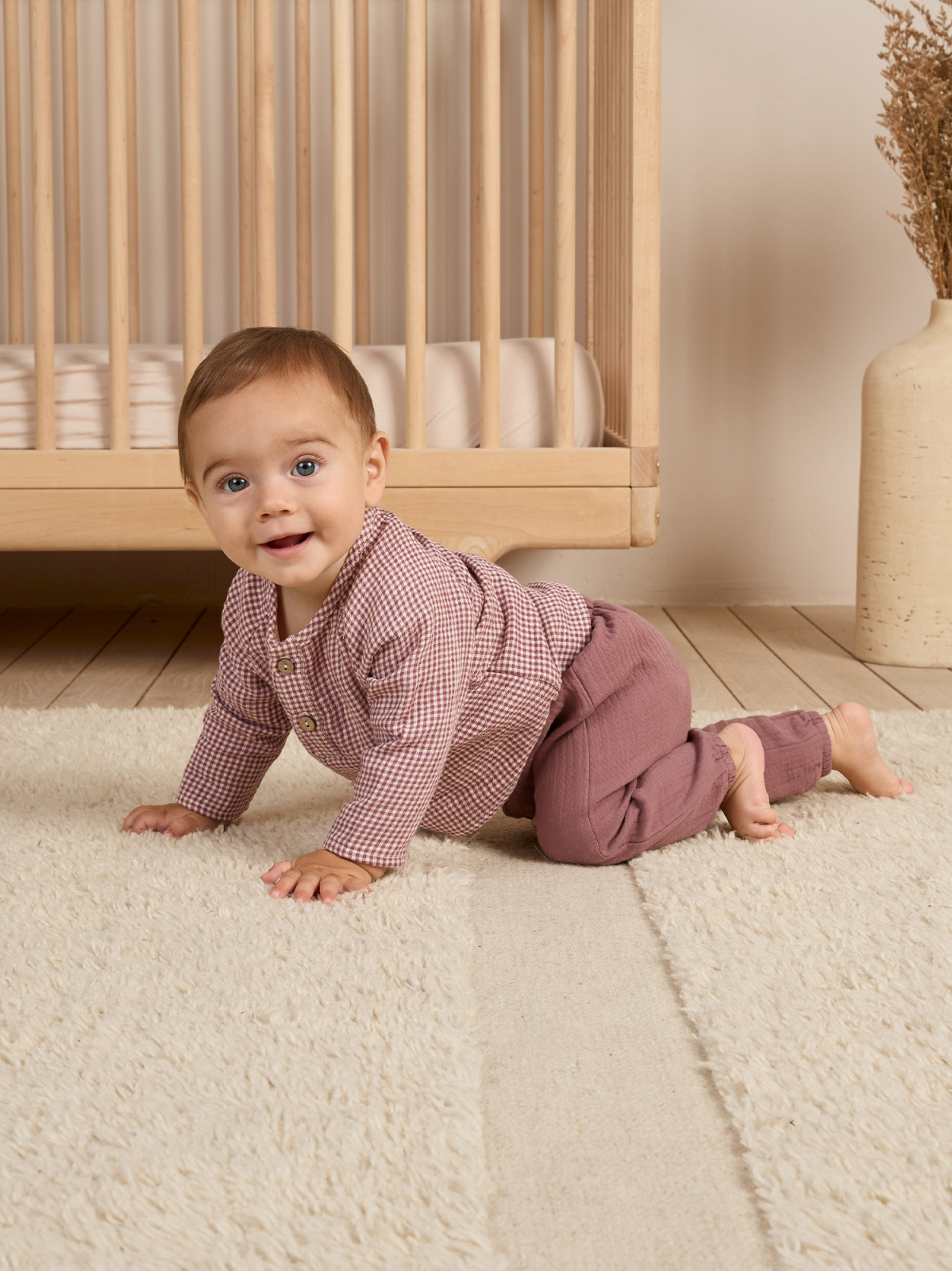 Smiling baby crawling on a fluffy rug in front of a wooden crib, wearing a checkered shirt and pants.