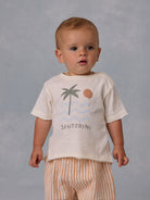 Toddler wearing a cream shirt with palm tree and sun design, striped pants, against a soft gray background.