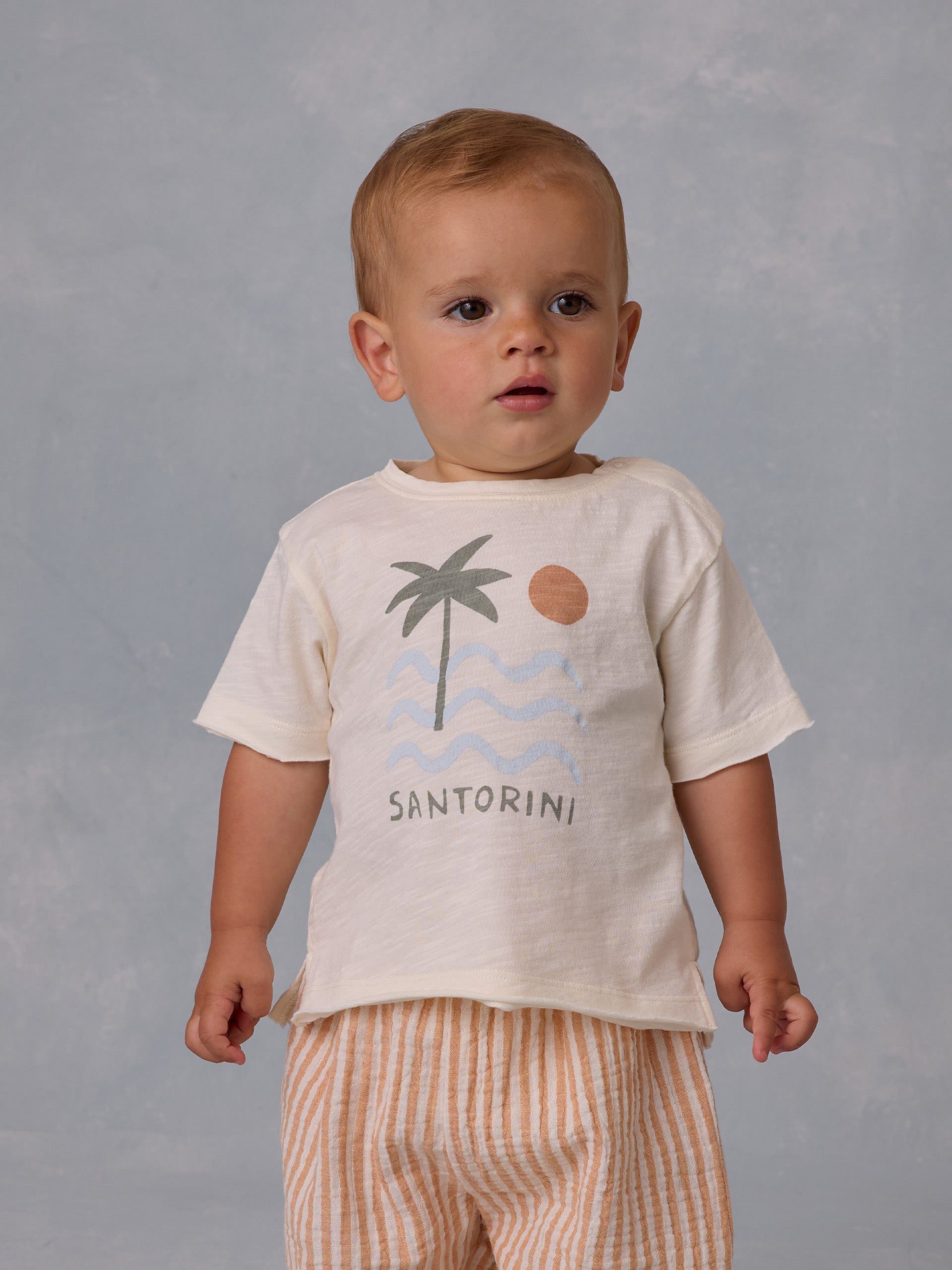 Toddler wearing a cream shirt with palm tree and sun design, striped pants, against a soft gray background.