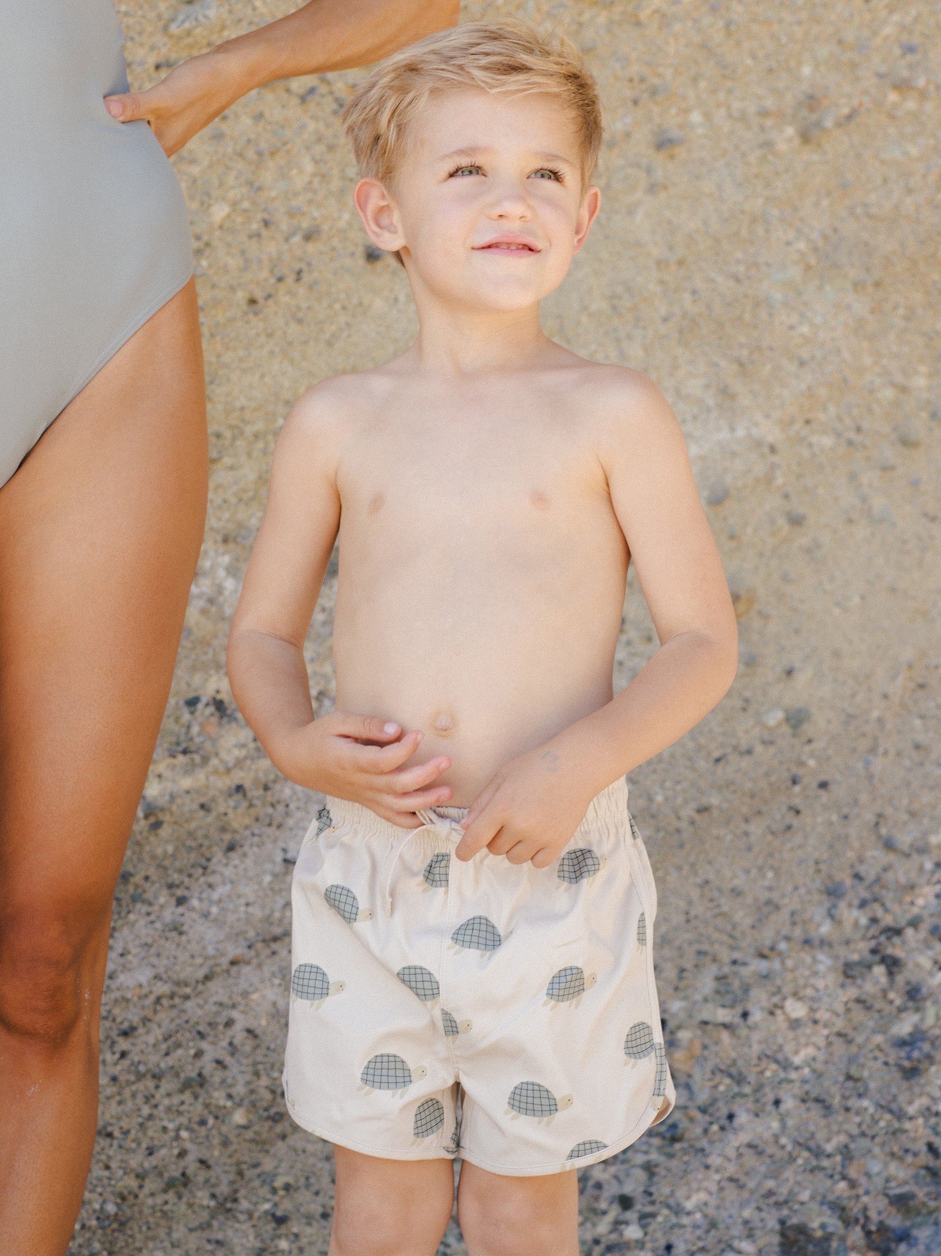Boy in turtle-pattern swim shorts smiles beside an adult in a swimsuit against a textured sandy background.