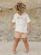 Child with curly hair wearing a "Santorini" shirt and palm-patterned shorts, playing in sandy beach setting.