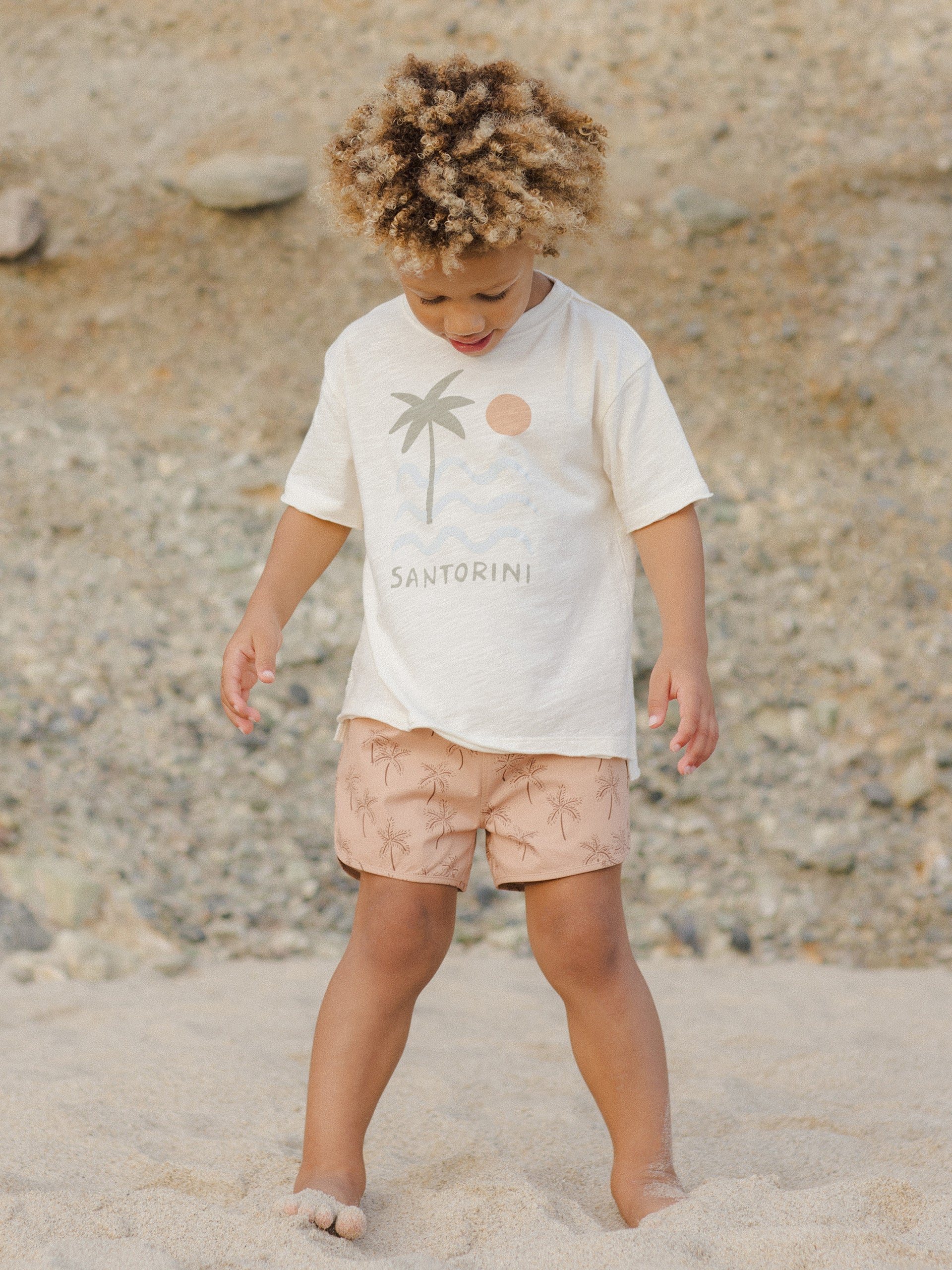 Child with curly hair wearing a "Santorini" shirt and palm-patterned shorts, playing in sandy beach setting.
