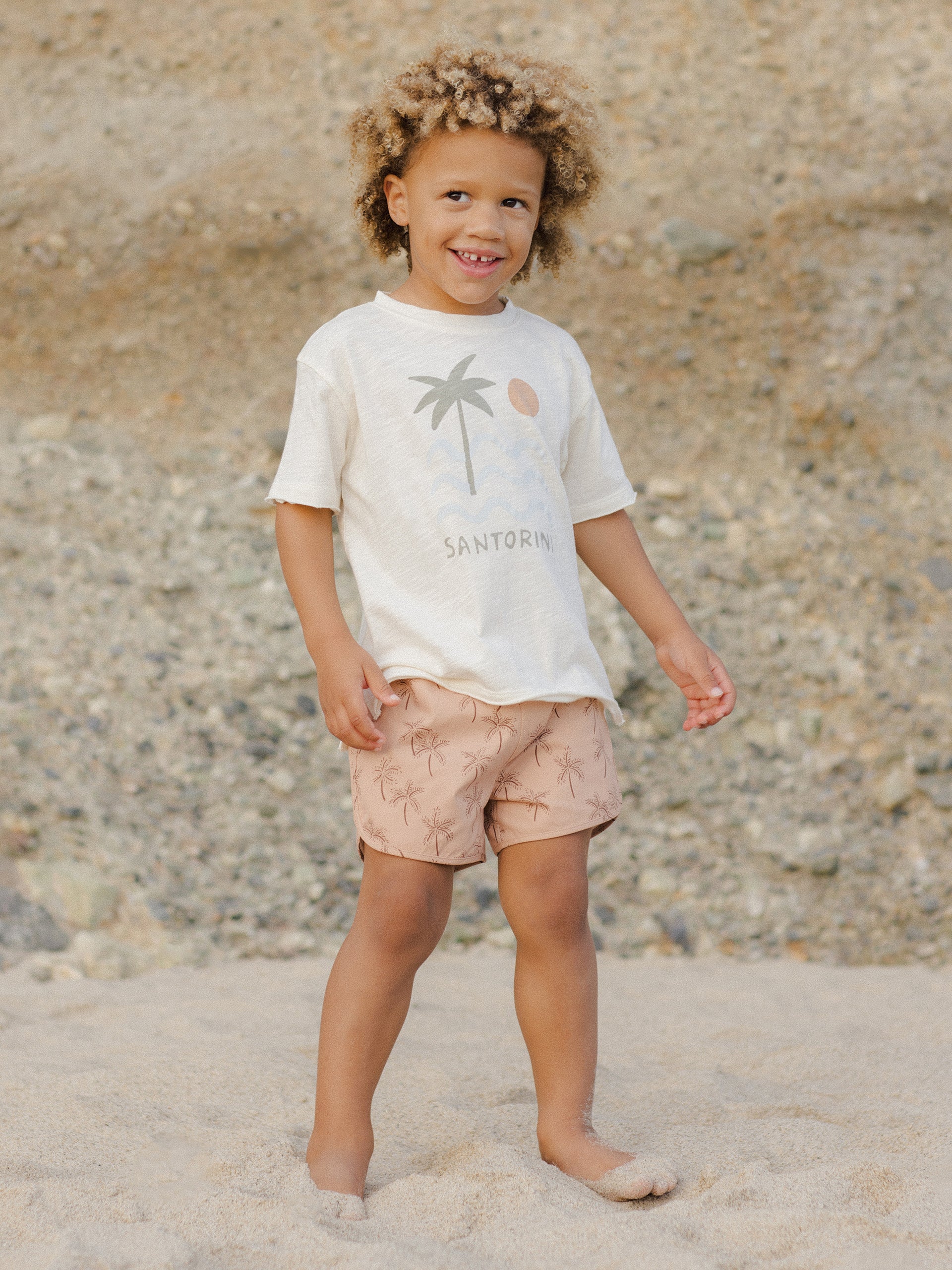 Child with curly hair in a light shirt and pink shorts, standing on sand with a rocky background.