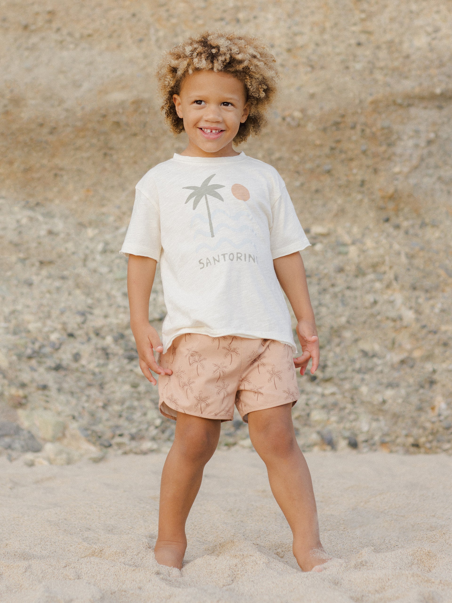 Smiling child with curly hair wearing a "Santorini" shirt and palm-patterned shorts on a beach.