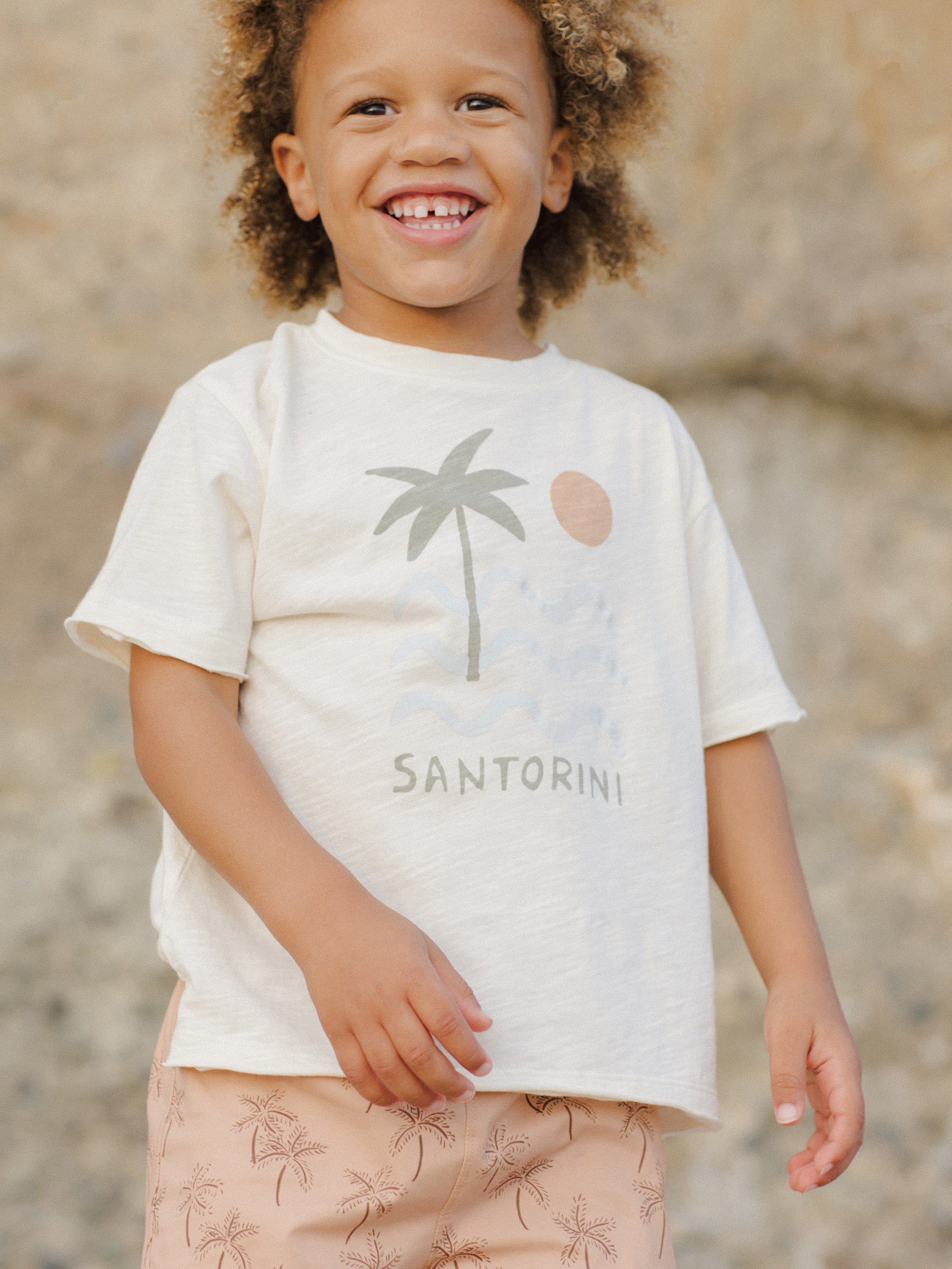 Smiling child in a light shirt with a palm tree graphic, standing outdoors against a rocky background.