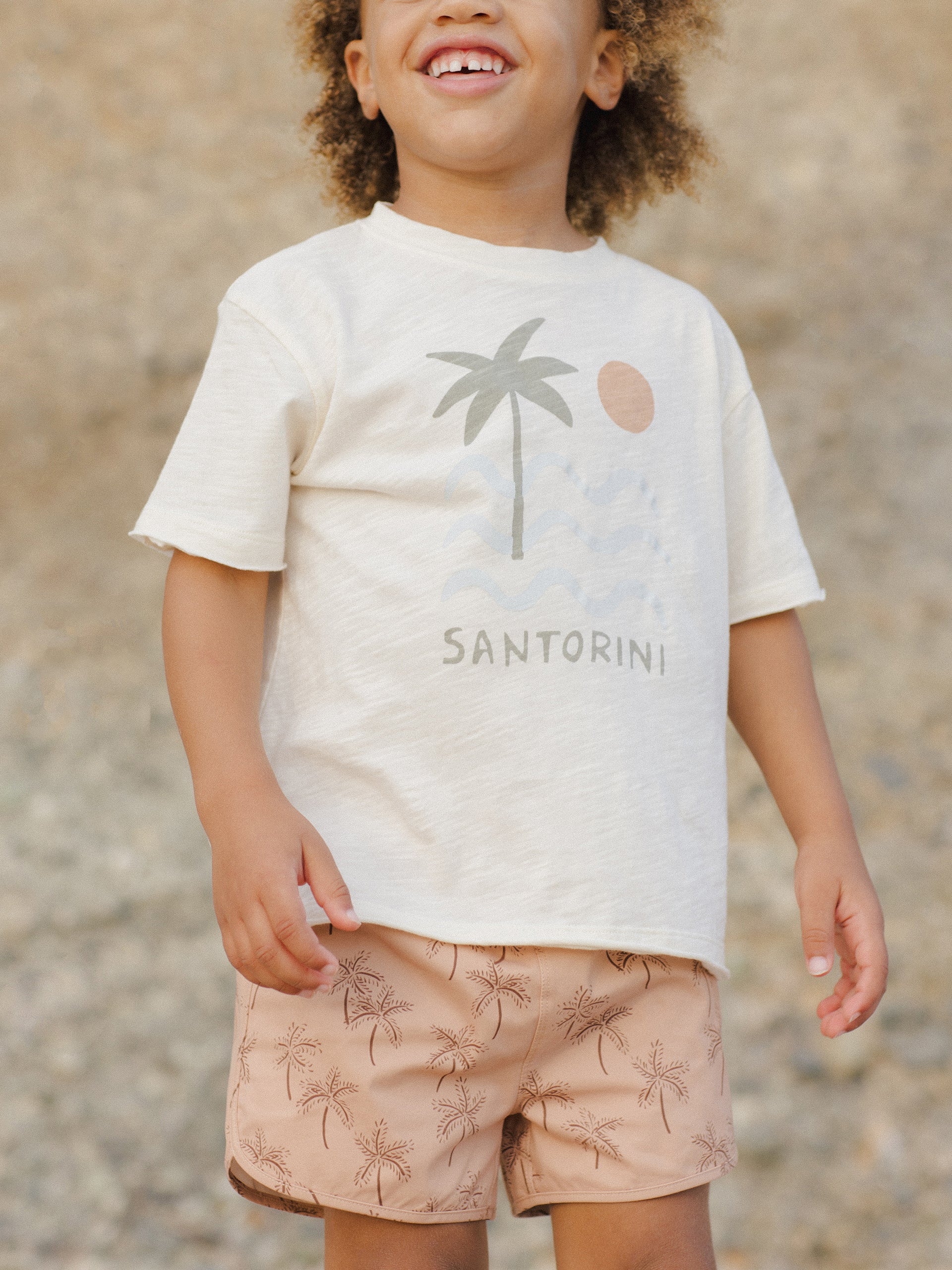 Child smiling in a palm tree graphic tee and patterned shorts, standing against a blurred natural background.