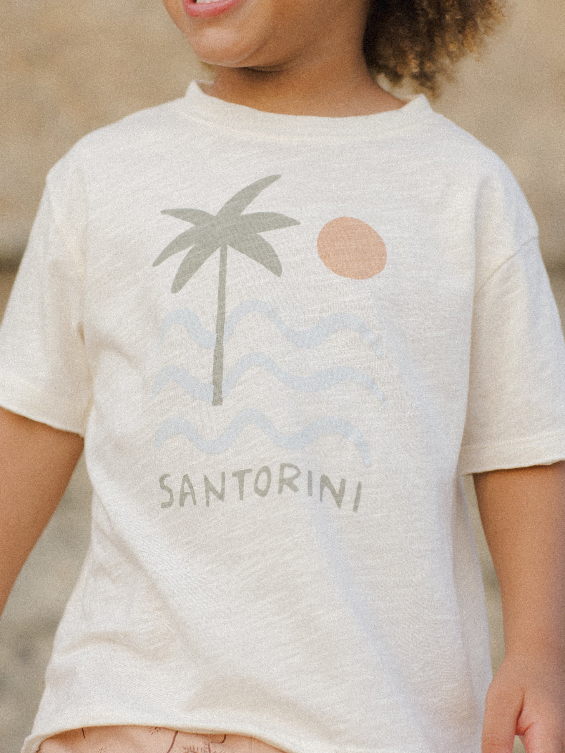 Child smiling while wearing a light shirt featuring a palm tree, waves, and a sun graphic labeled "Santorini."