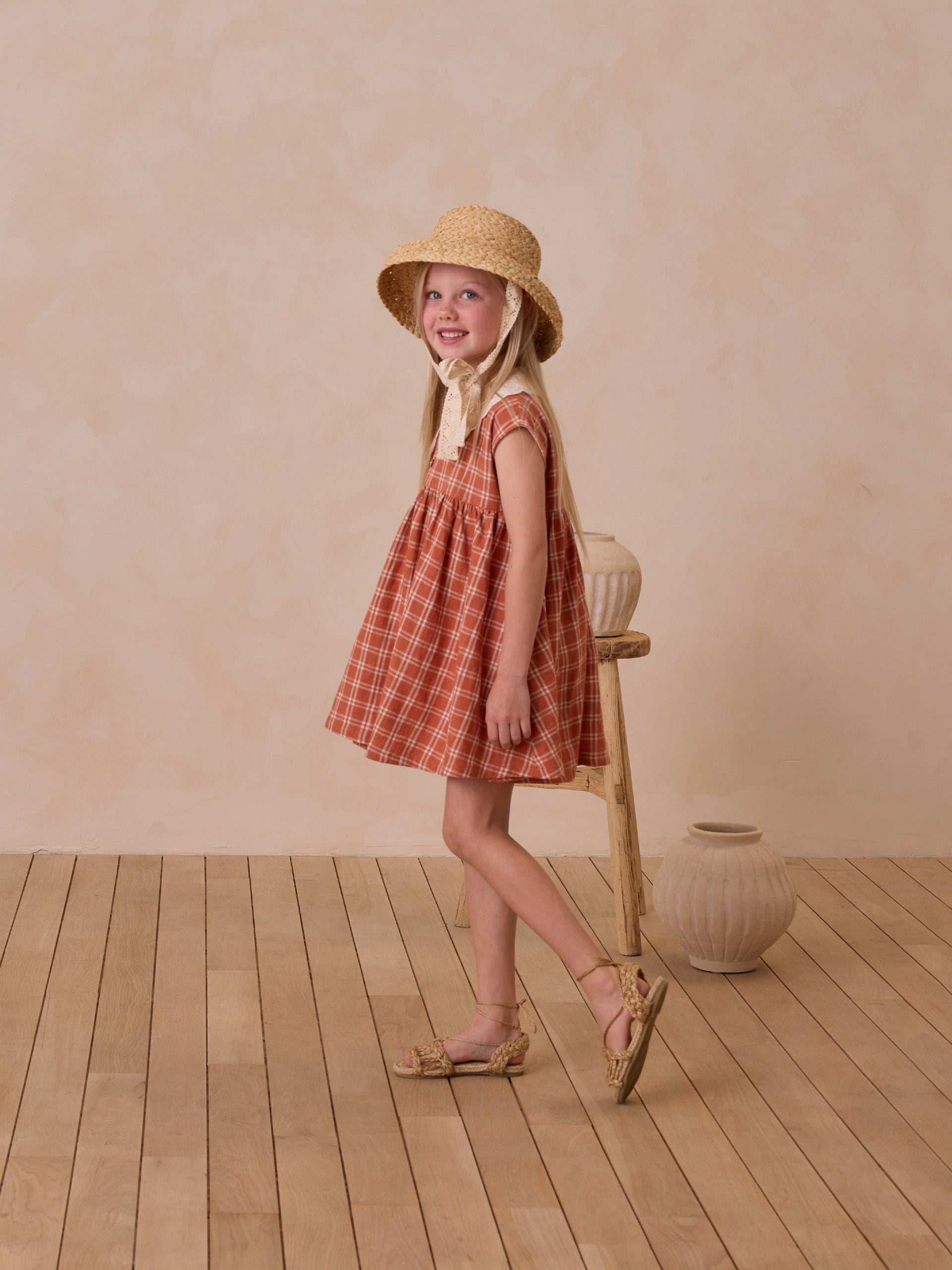 Smiling girl in a plaid dress and straw hat, standing beside a wooden stool and clay pots on a wooden floor.