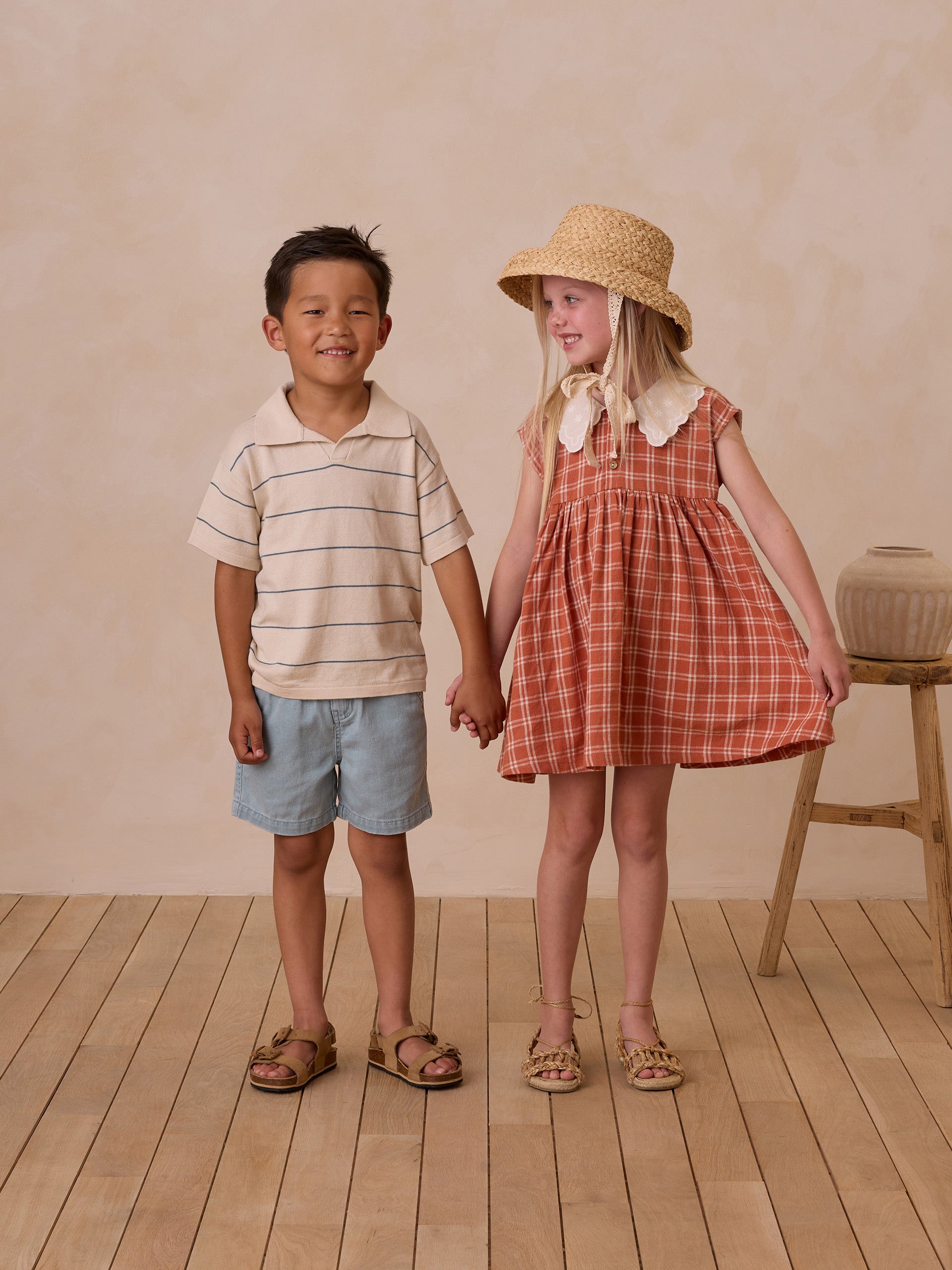 Two children holding hands, smiling. A boy in a striped shirt and shorts, and a girl in a plaid dress with a straw hat.
