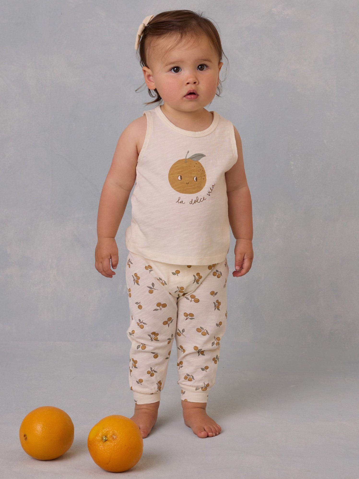 Toddler in a fruit-themed outfit stands beside oranges on a soft surface, looking curiously at the camera.