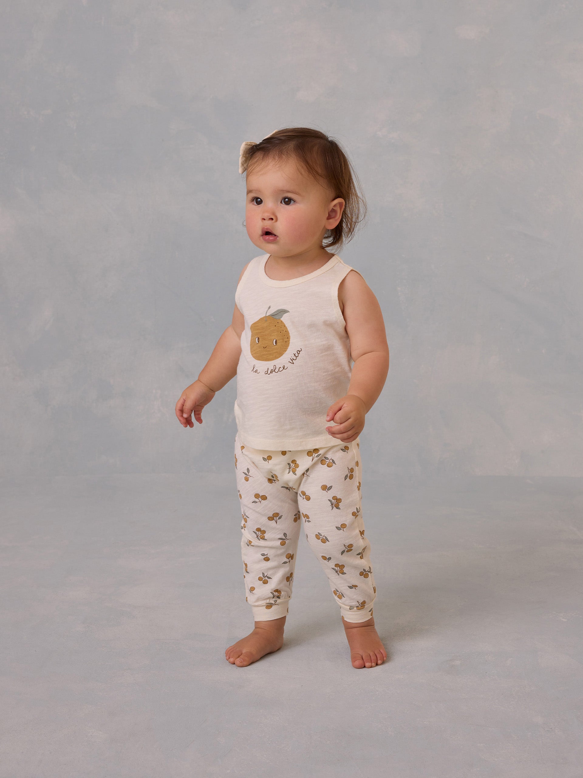 Toddler wearing a tank top with an orange graphic and patterned pants, standing on a light textured background.