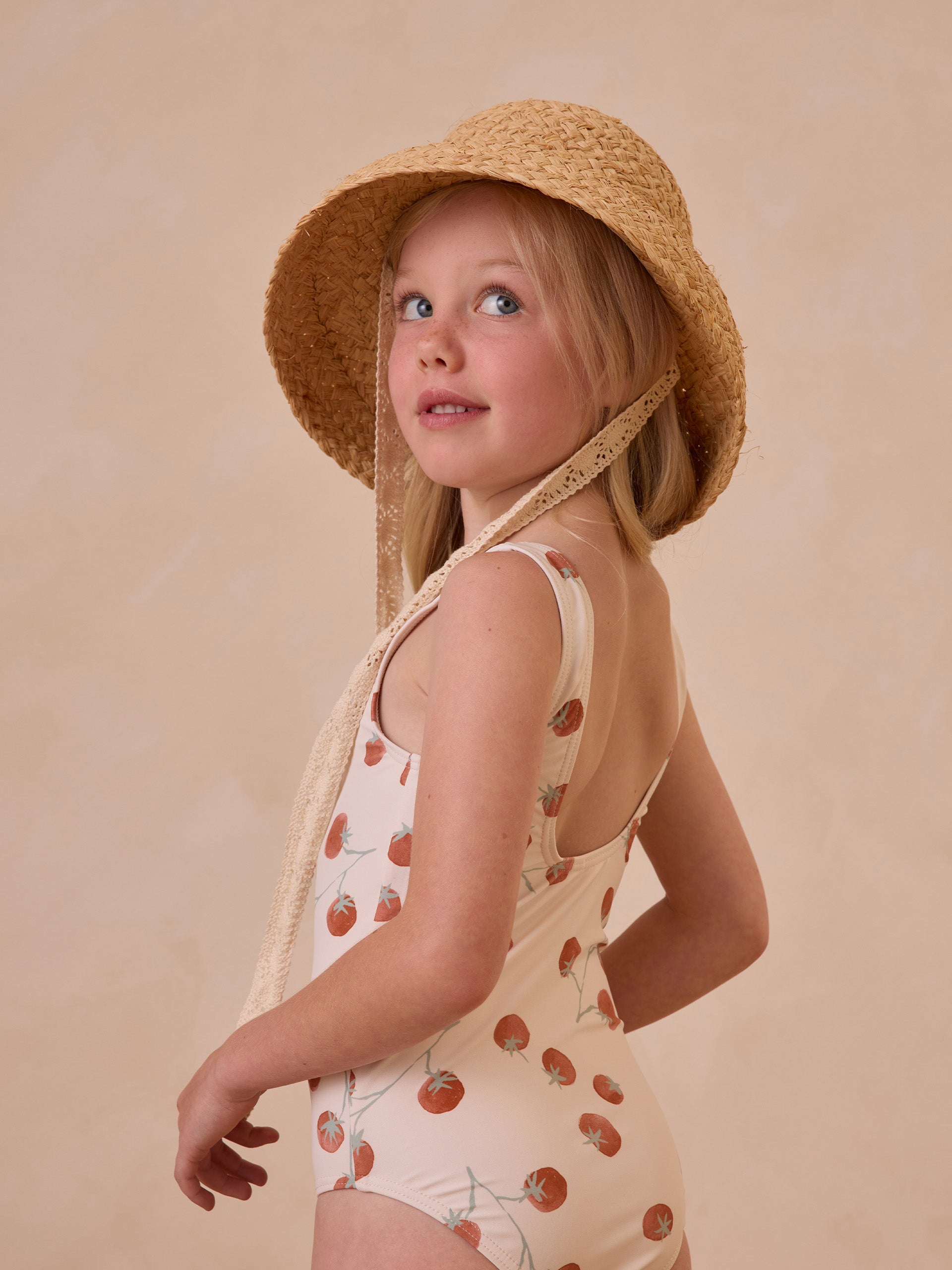 Girl in a floral swimsuit and straw hat, looking over her shoulder, with a beige background.