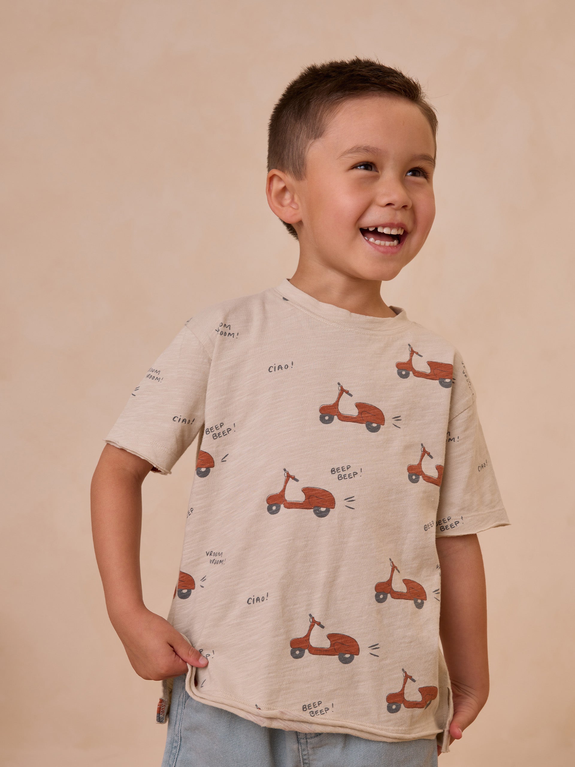 Smiling boy in a light t-shirt adorned with scooter graphics against a soft, neutral background.
