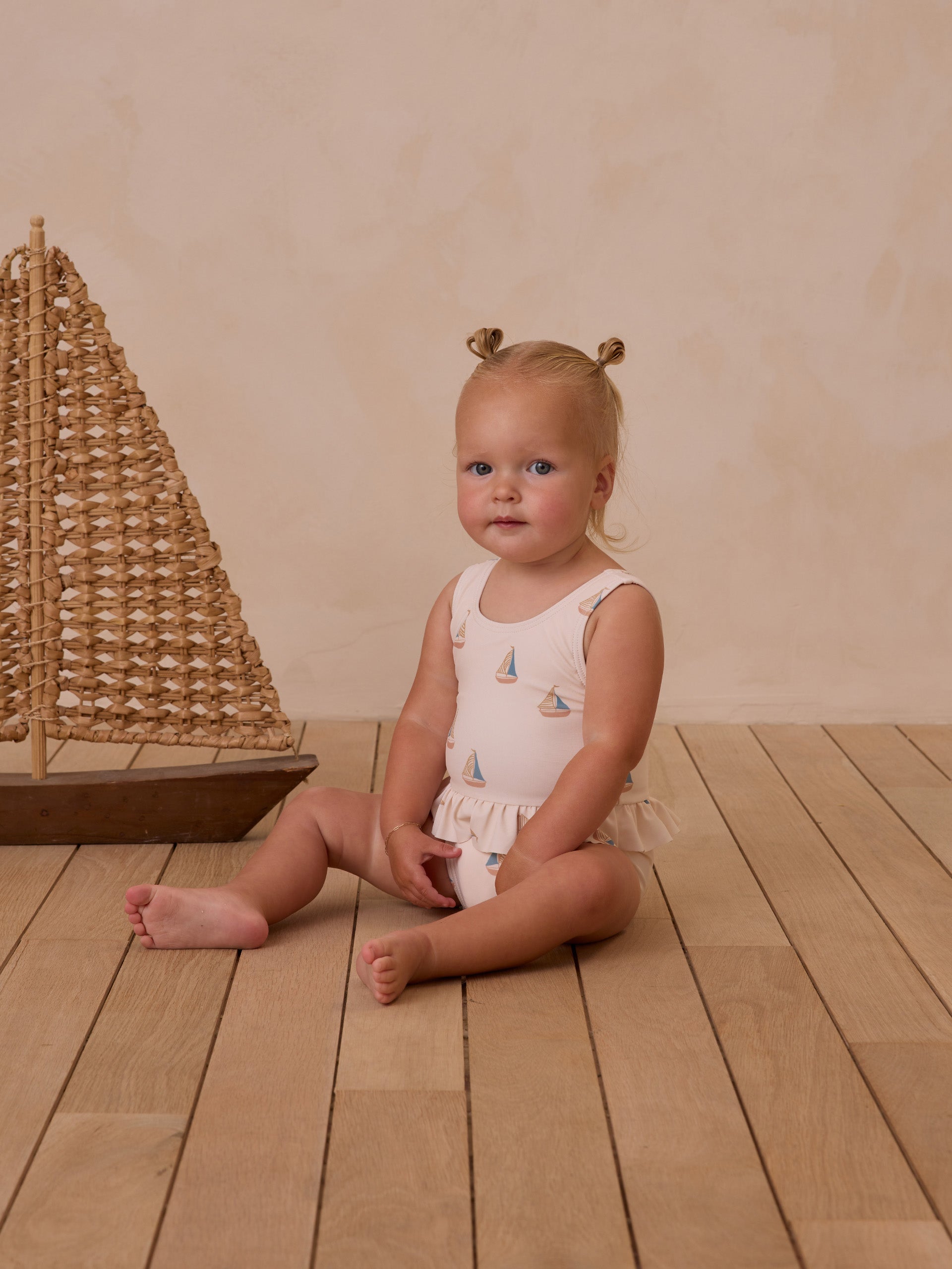 Toddler girl sits on wooden floor beside a decorative sailboat, wearing a light-colored swimsuit with sailprints.