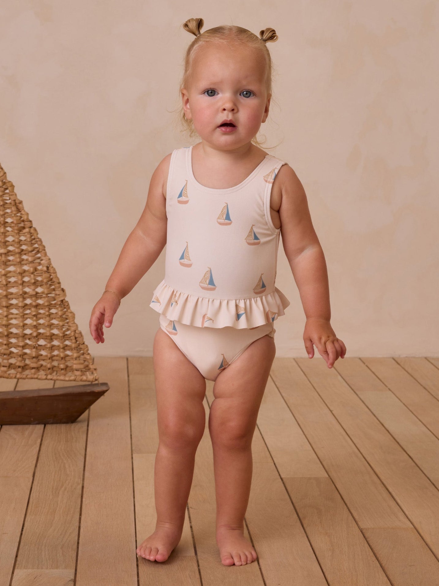 Toddler in a sailboat-patterned swimsuit with ruffled edges, standing on wooden floor against a beige background.