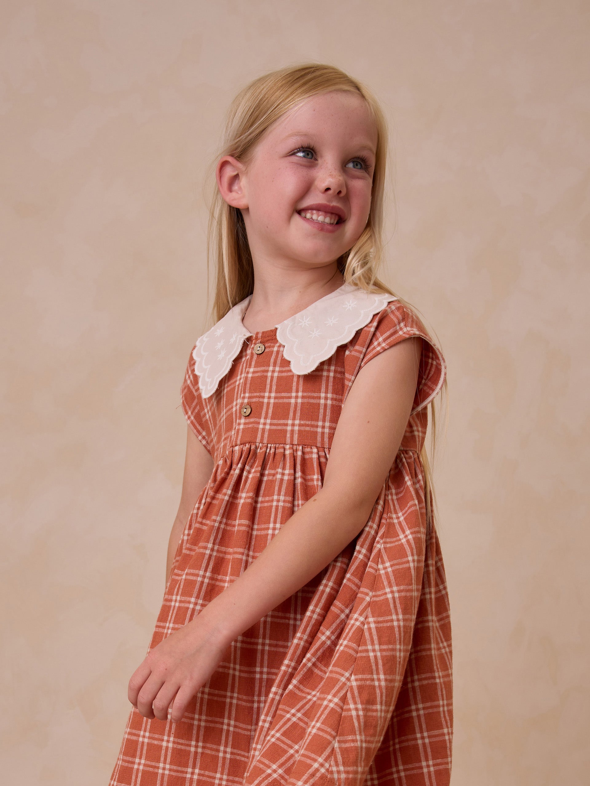 Smiling girl in a checked dress with a white collar, standing against a soft beige background.