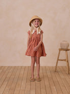 Smiling girl in a plaid dress and straw hat stands on wooden floor against a textured background.
