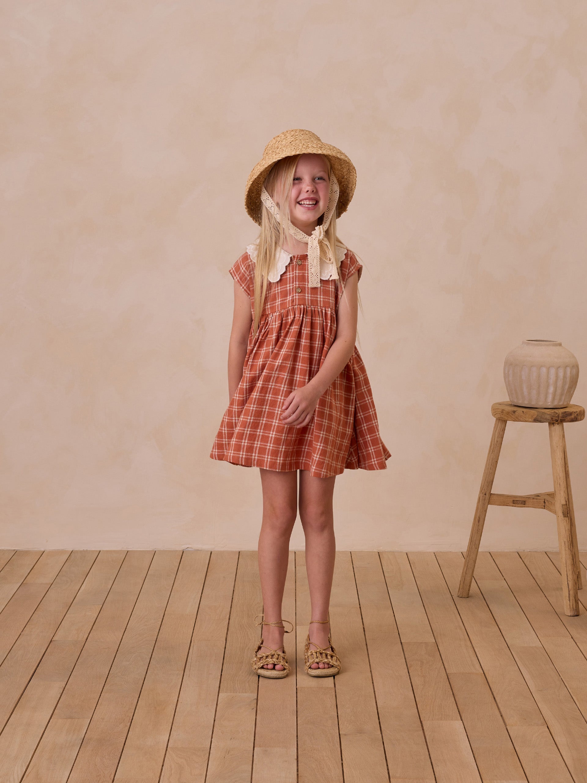 Smiling girl in a plaid dress and straw hat stands on wooden floor against a textured background.