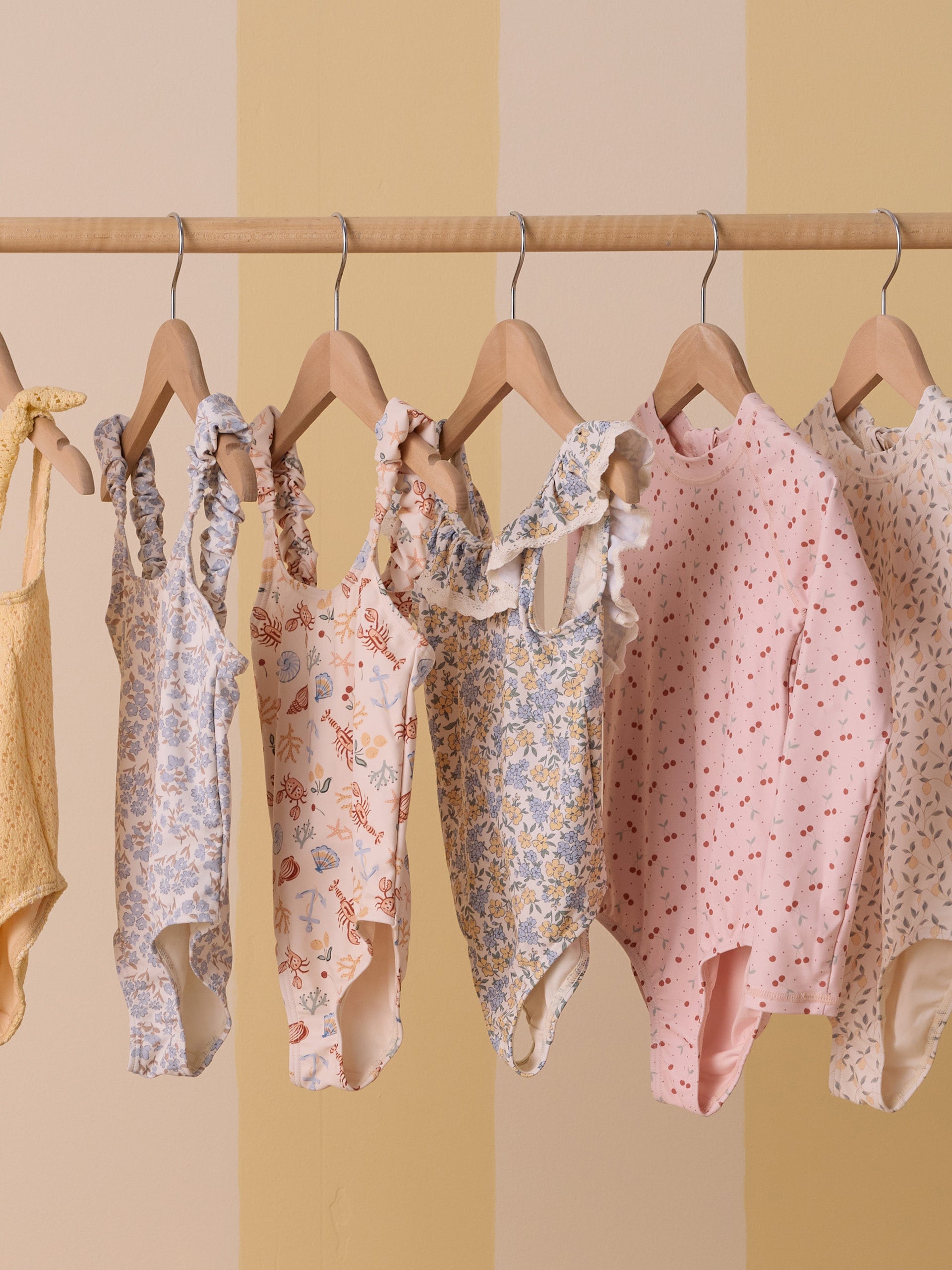 Colorful children's swimsuits hanging on a wooden rack against a soft yellow striped background.