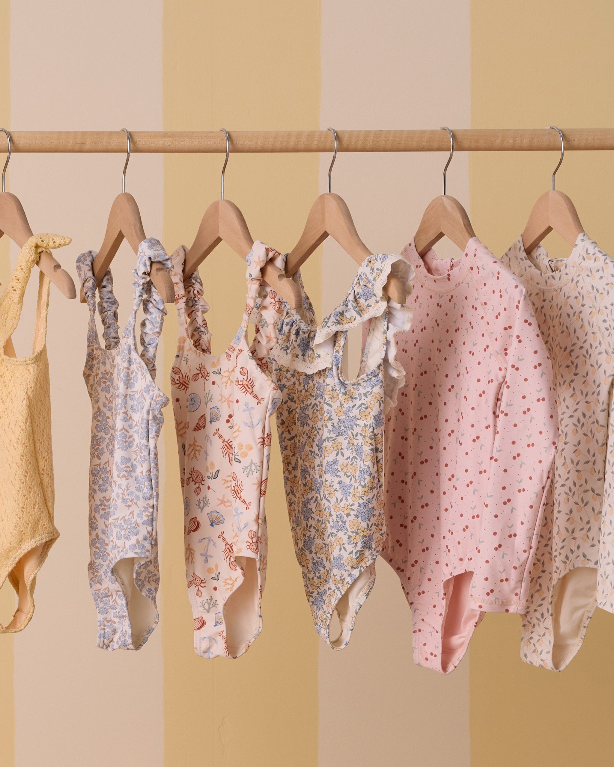 Colorful children's swimsuits hanging on a wooden rack against a soft yellow striped background.
