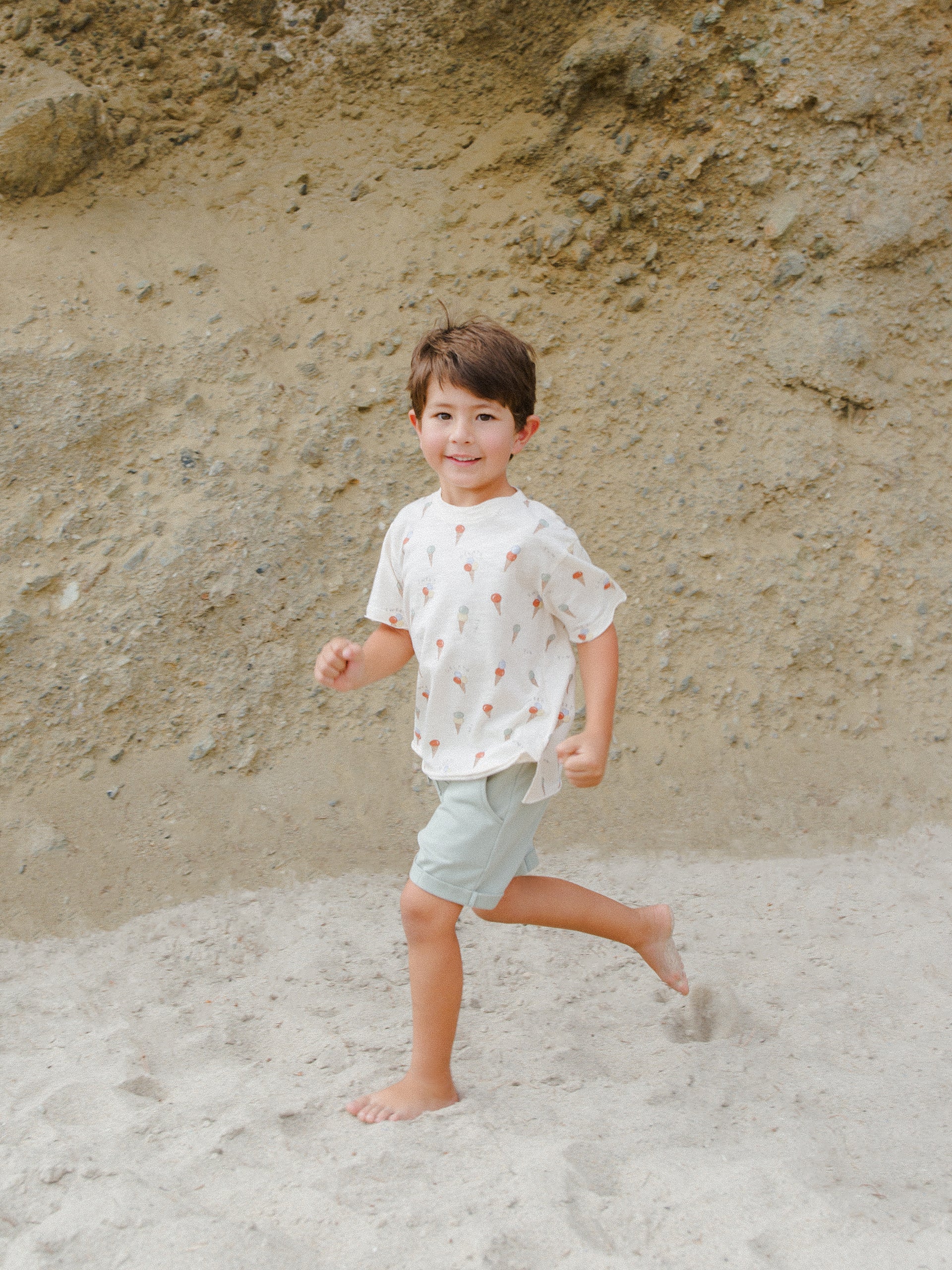 Smiling boy in a playful shirt running on a sandy beach with a rocky background.
