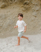 Smiling boy in a playful shirt running on a sandy beach with a rocky background.