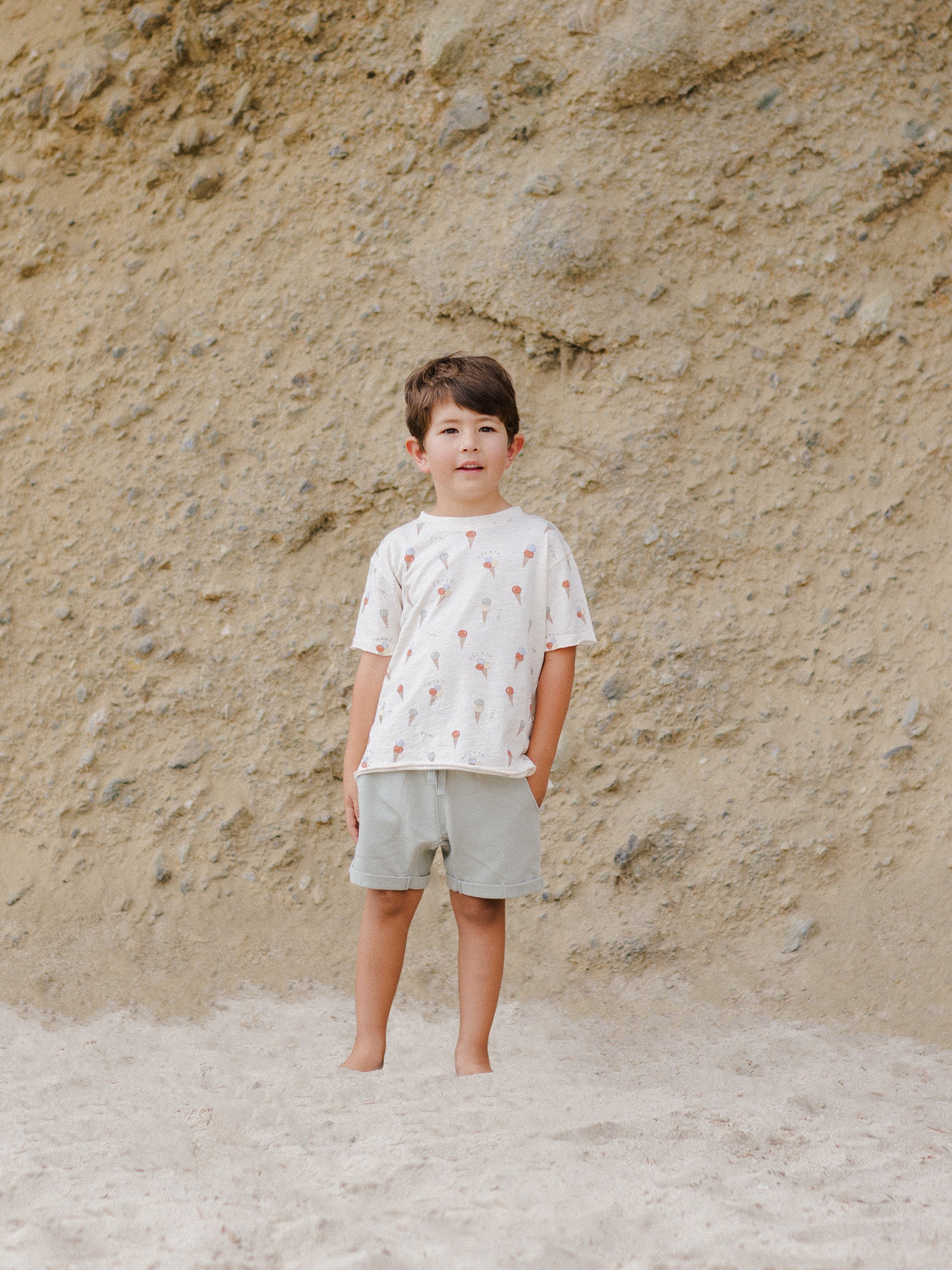 Child standing on sandy ground wearing a patterned shirt and shorts, with a rocky background.