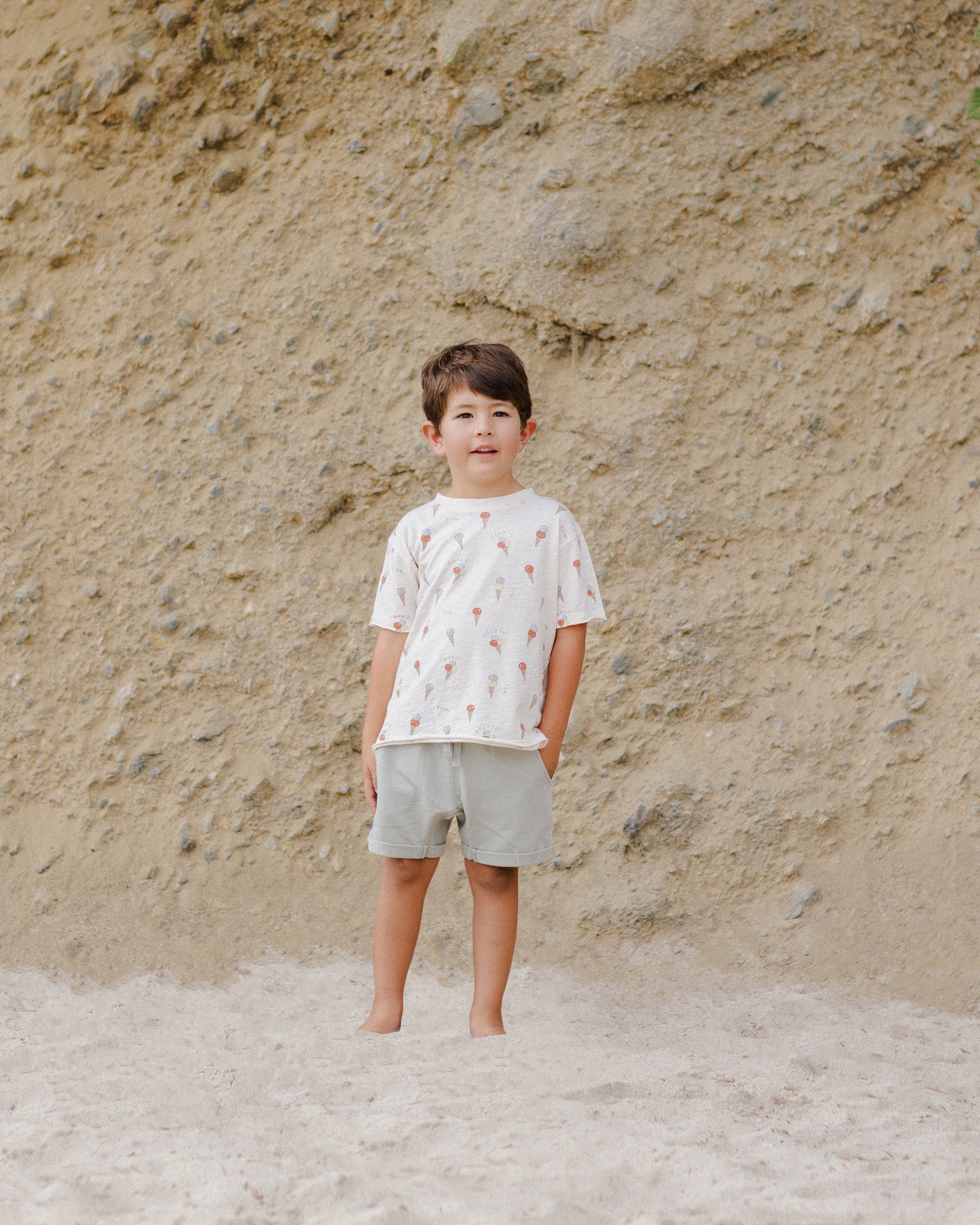 Child standing on sandy ground wearing a patterned shirt and shorts, with a rocky background.