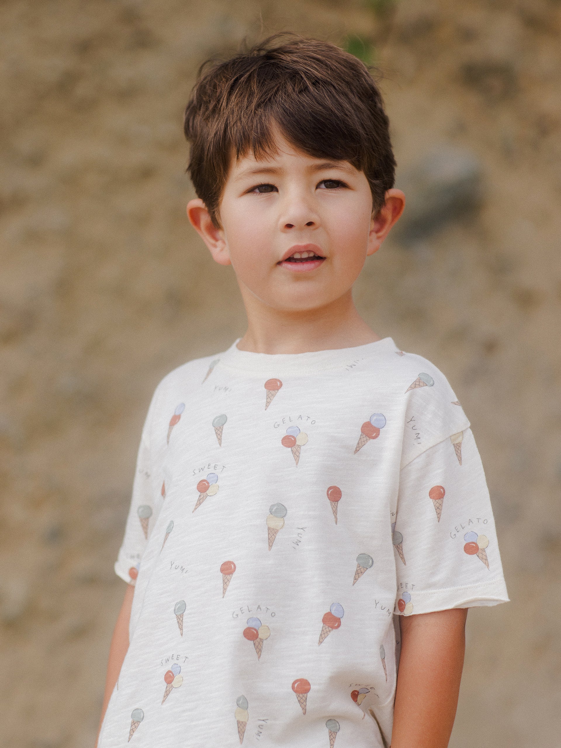 Child in a gelato-patterned shirt stands outdoors, looking thoughtfully at the camera against a natural background.