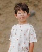 Child in a gelato-patterned shirt stands outdoors, looking thoughtfully at the camera against a natural background.
