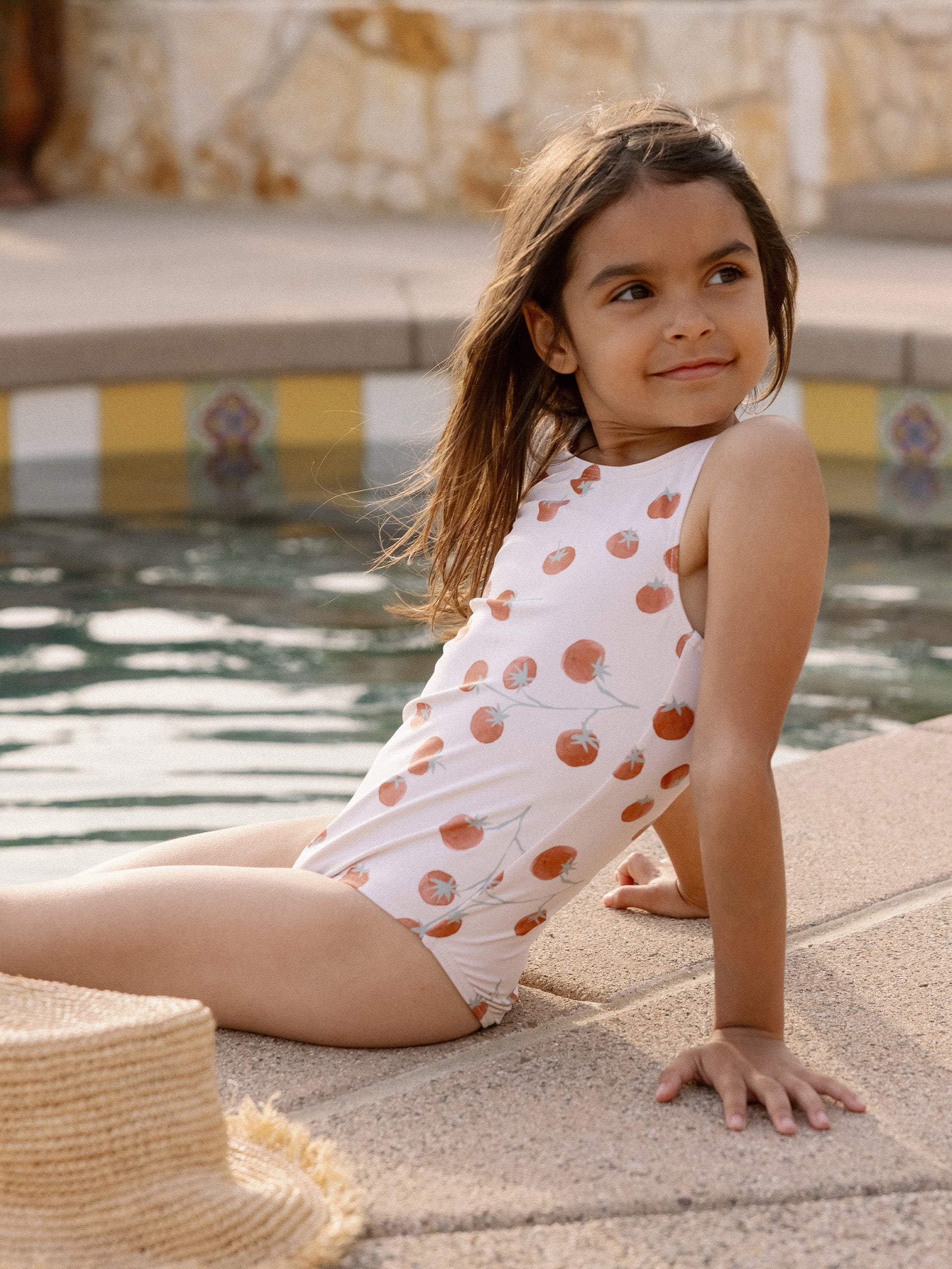 Girl in a swimsuit with a tomato print, sitting by a pool, smiling at the camera on a sunny day.