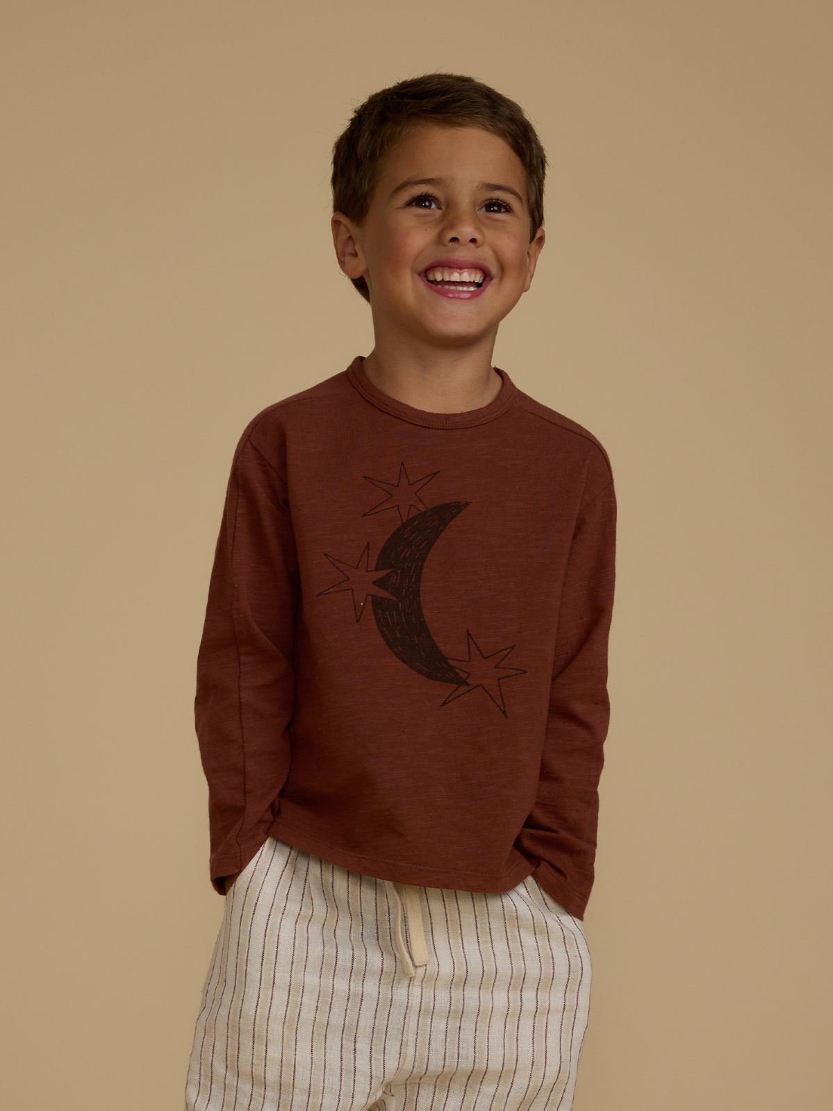 Smiling child in a brown long-sleeve shirt with moon and stars design, wearing light striped shorts against a beige background.