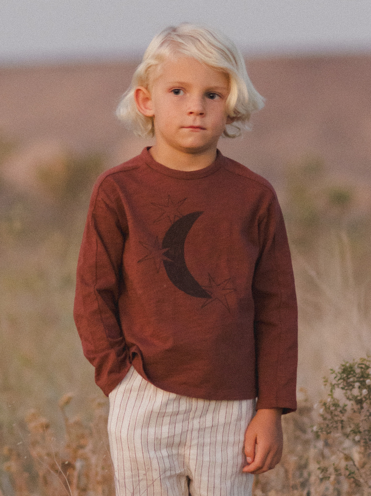 Child in a brown long-sleeve shirt with moon design, standing in a grassy field during golden hour.