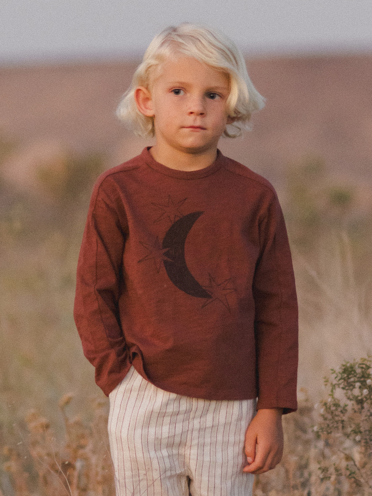 Child in a brown long-sleeve shirt with moon design, standing in a grassy field during golden hour.