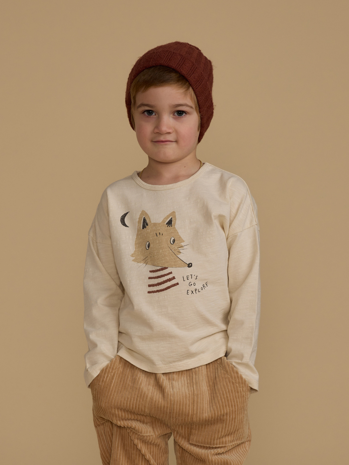 Child wearing a beige shirt with a fox graphic and striped hem, paired with corduroy pants and a brown beanie.
