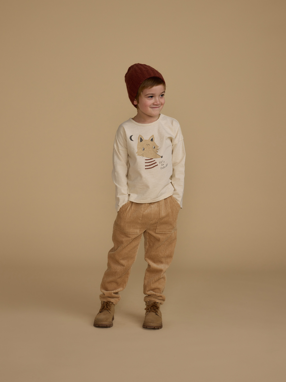 Child wearing a beige shirt with a fox design, tan corduroy pants, and a brown knit hat against a neutral background.